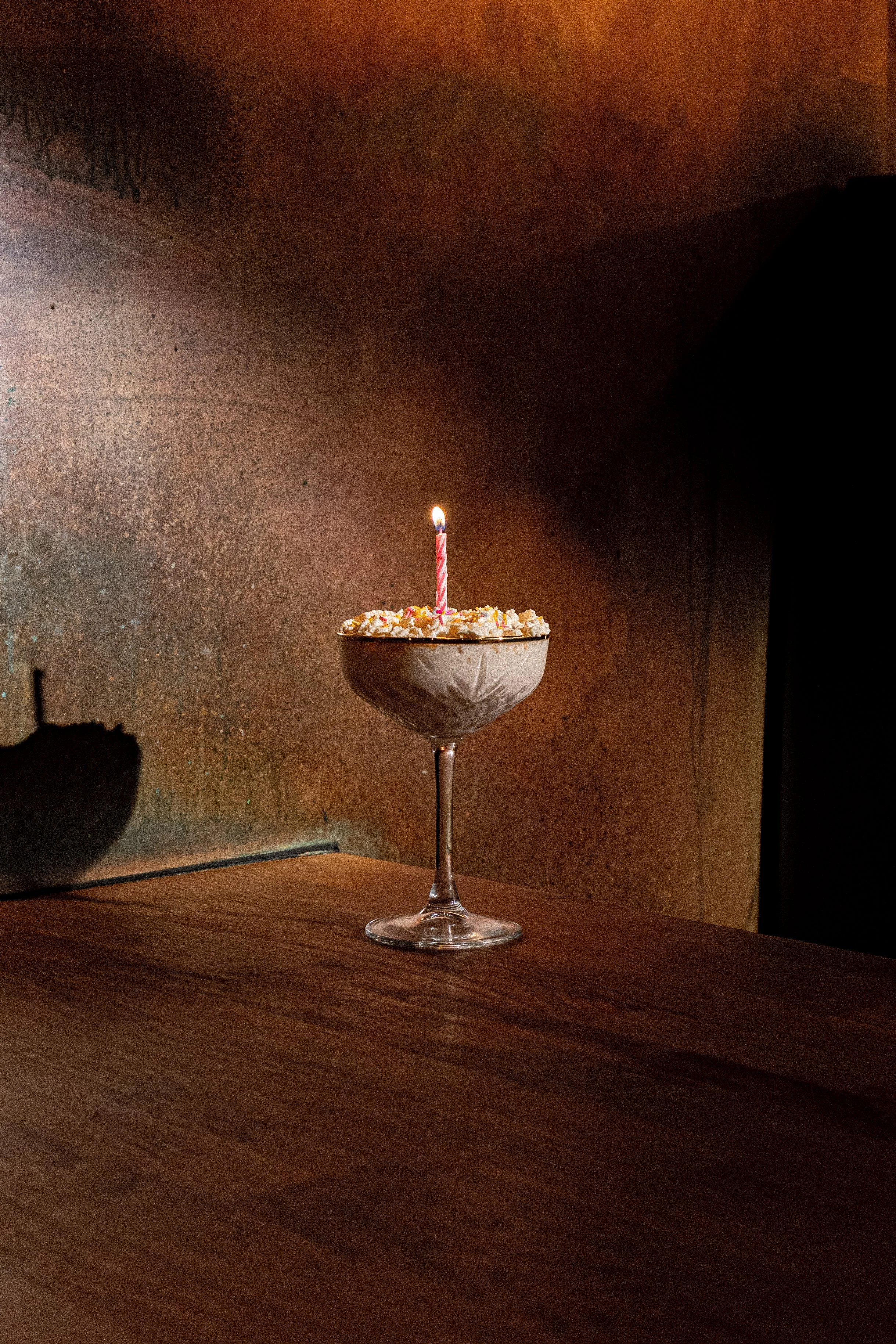 A dessert in a martini glass with a lit birthday candle on top, placed on a wooden surface with a dark background.