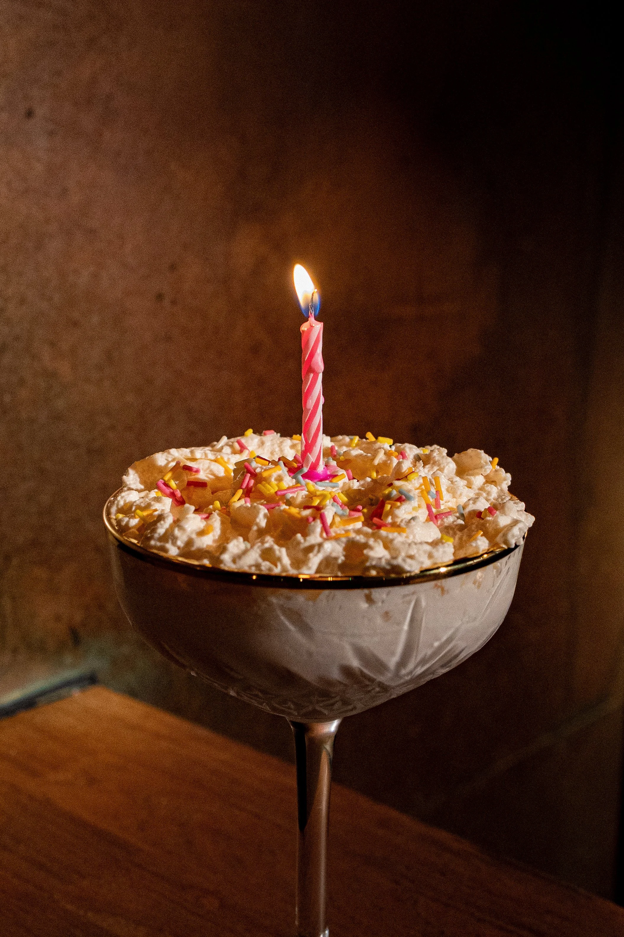A cocktail glass filled with whipped cream and colorful sprinkles, topped with a lit pink birthday candle.