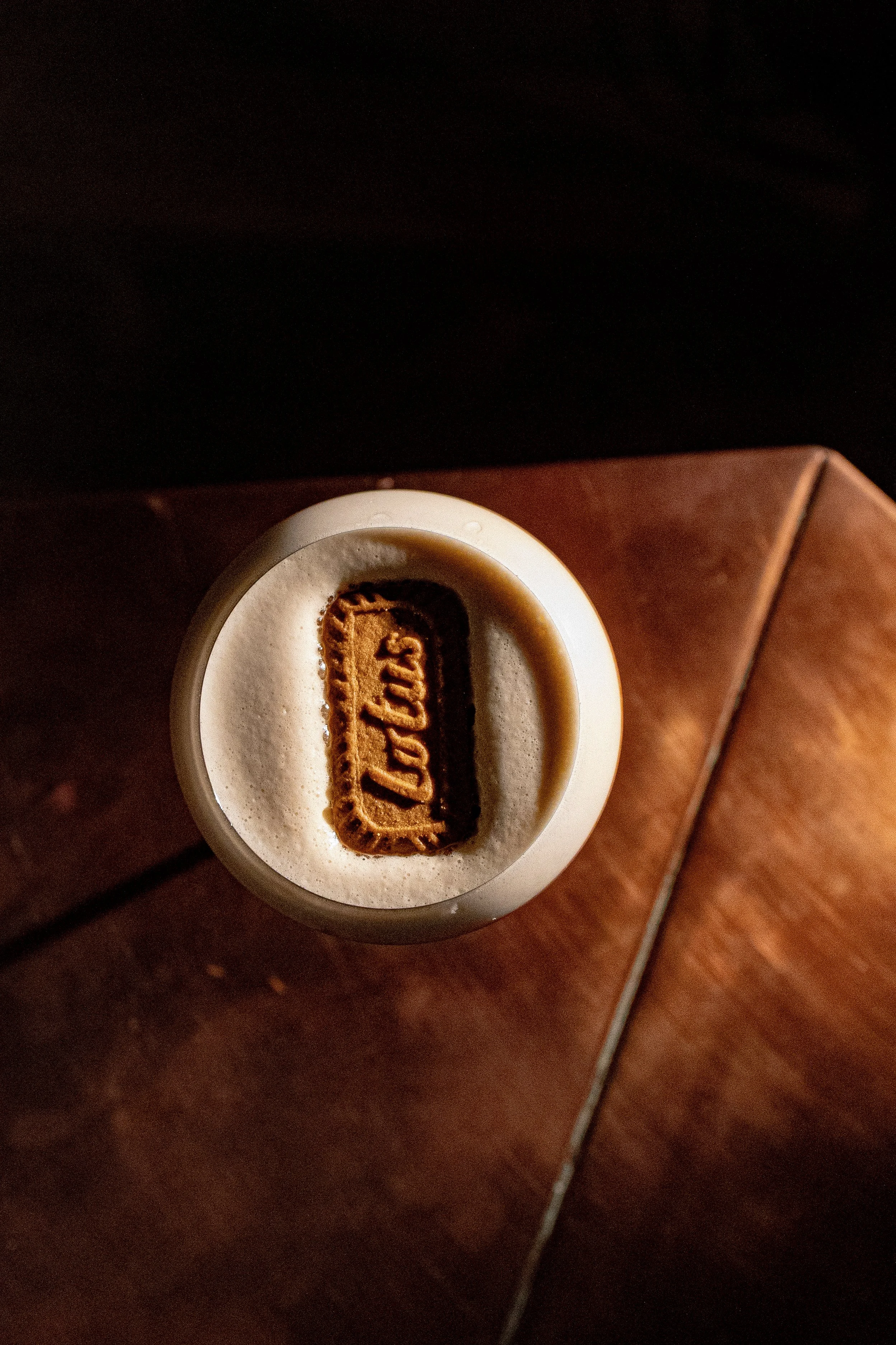 A white coffee mug filled with coffee, topped with foam and a Lotus biscuit on top, placed on a wooden surface.