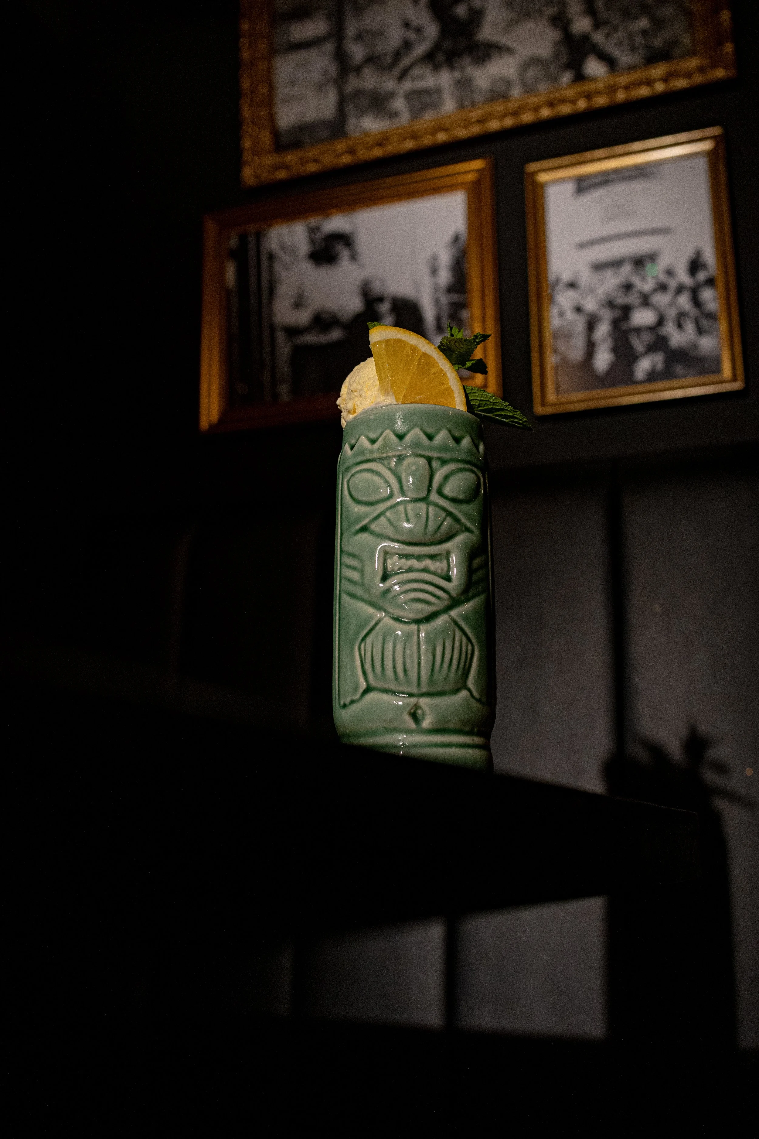 A tiki-style ceramic mug with a face design holding a lemon wedge, mint leaves, and a scoop of ice cream, placed on a dark table against a dimly lit background with framed black-and-white photographs.
