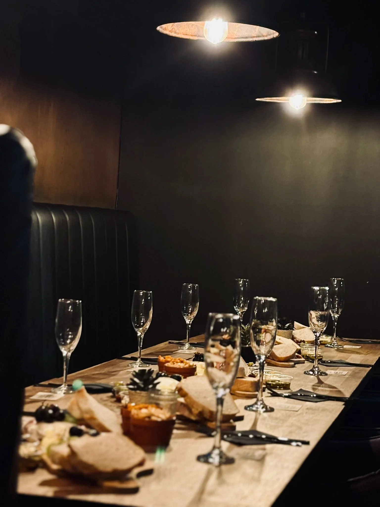 A dimly lit wooden dining table set with champagne glasses, sliced bread, small bowls of appetizers, and opened jars, with two hanging light fixtures casting warm light.