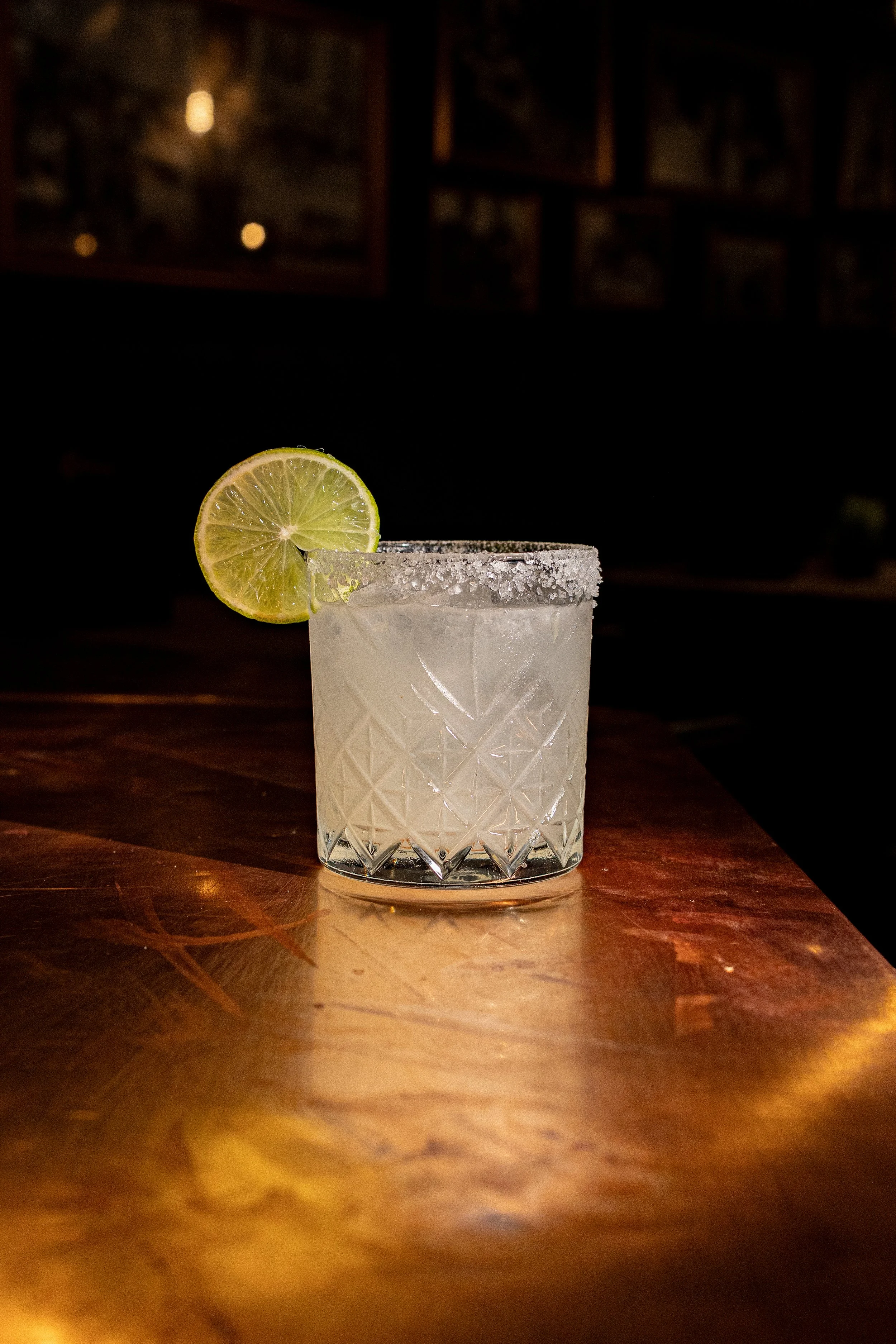 A glass of a light-colored cocktail garnished with a lime slice on a salted rim, placed on a wooden table in a dimly lit bar setting.