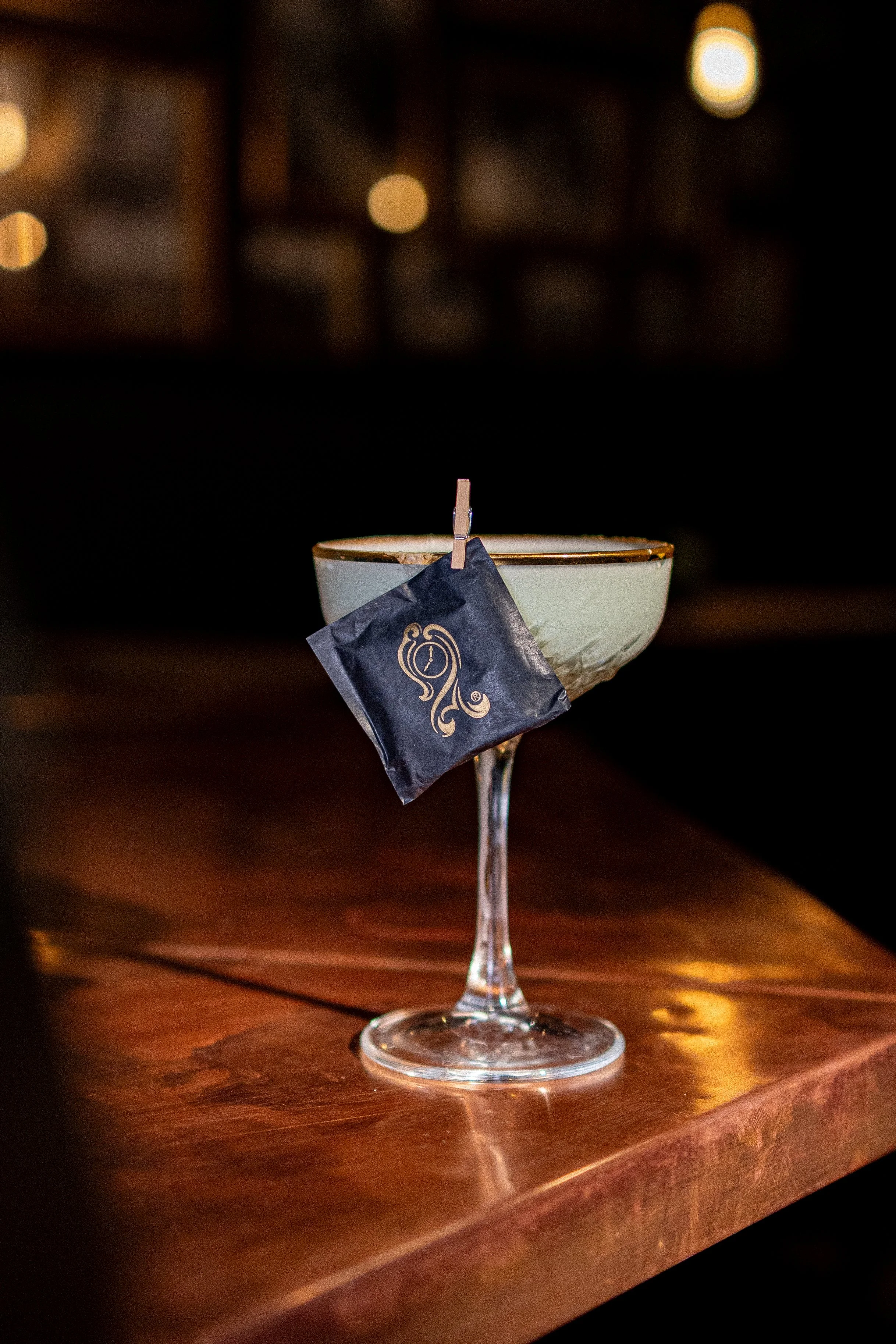 Cocktail glass with a tea bag hanging on the rim, placed on a wooden bar counter.