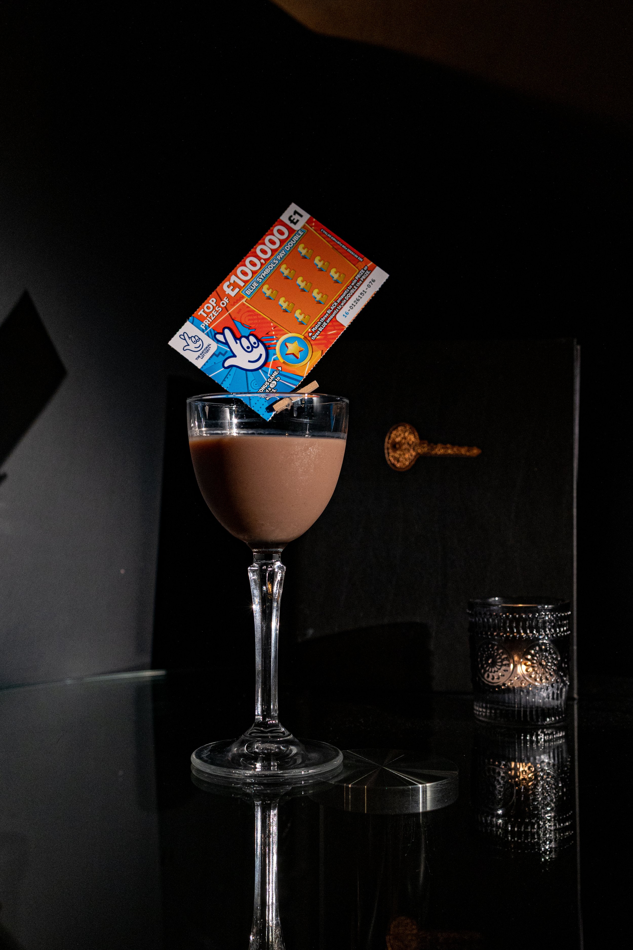 Glass of chocolate cocktail with a lottery scratcher ticket inserted at an angle, placed on a reflective black surface with a lit candle holder in the background.