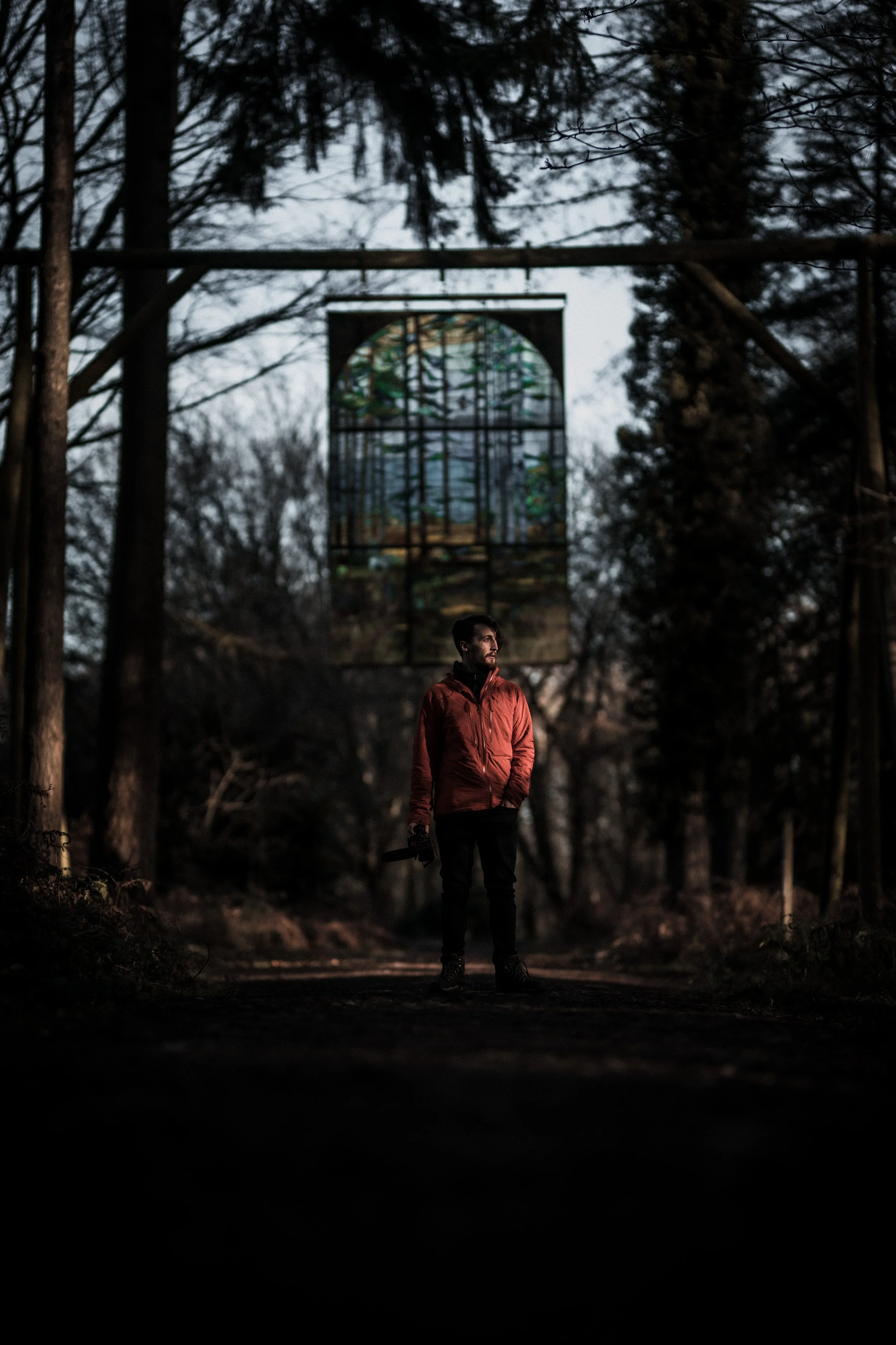 A man in an orange jacket holding a camera stands on a dark forest path with tall trees, and a stained glass window hangs above.