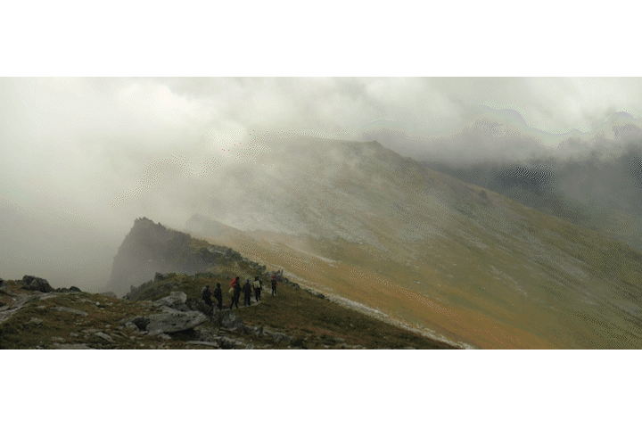 Group of hikers walking along a mountain trail with misty clouds and fog-covered mountains in the background.