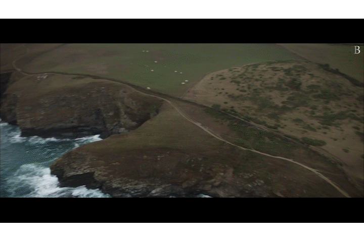 Aerial view of a rugged coastline with cliffs, a beach, and grassy fields, with a winding trail or road running through the landscape.
