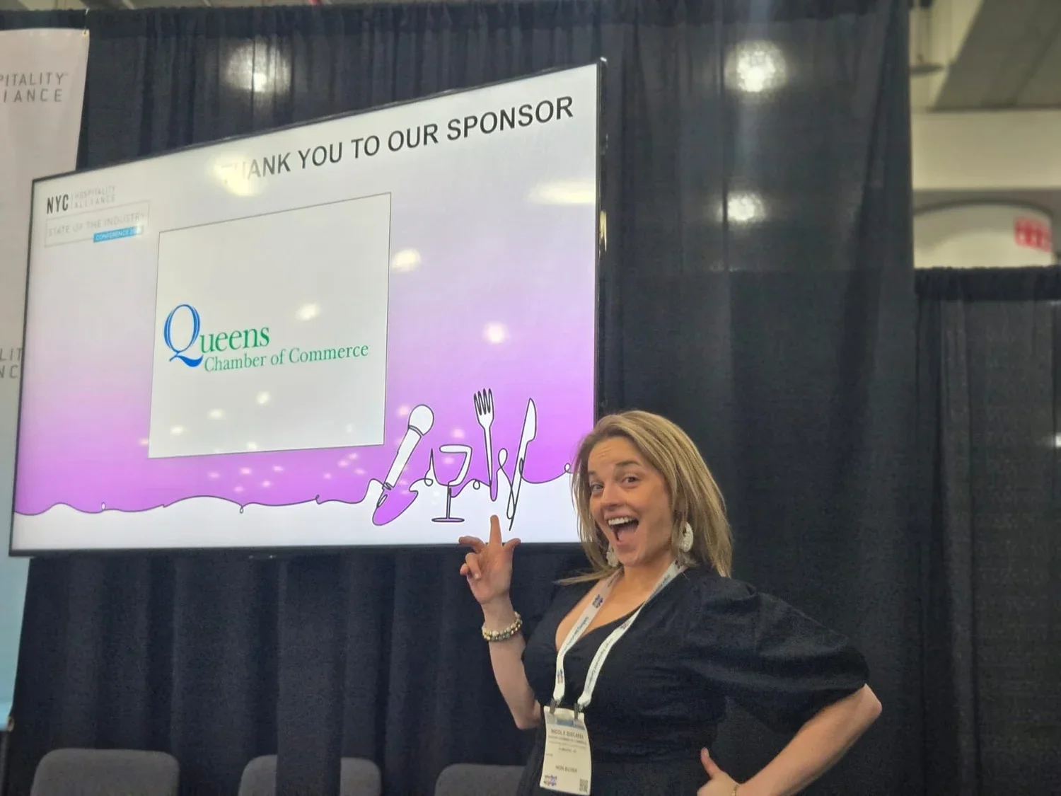 A woman standing in front of a large screen display at a conference, smiling and pointing at the screen which has a message of thanks to a sponsor, Queens Chamber of Commerce, with illustrations of a microphone, fork, knife, and glass of wine.