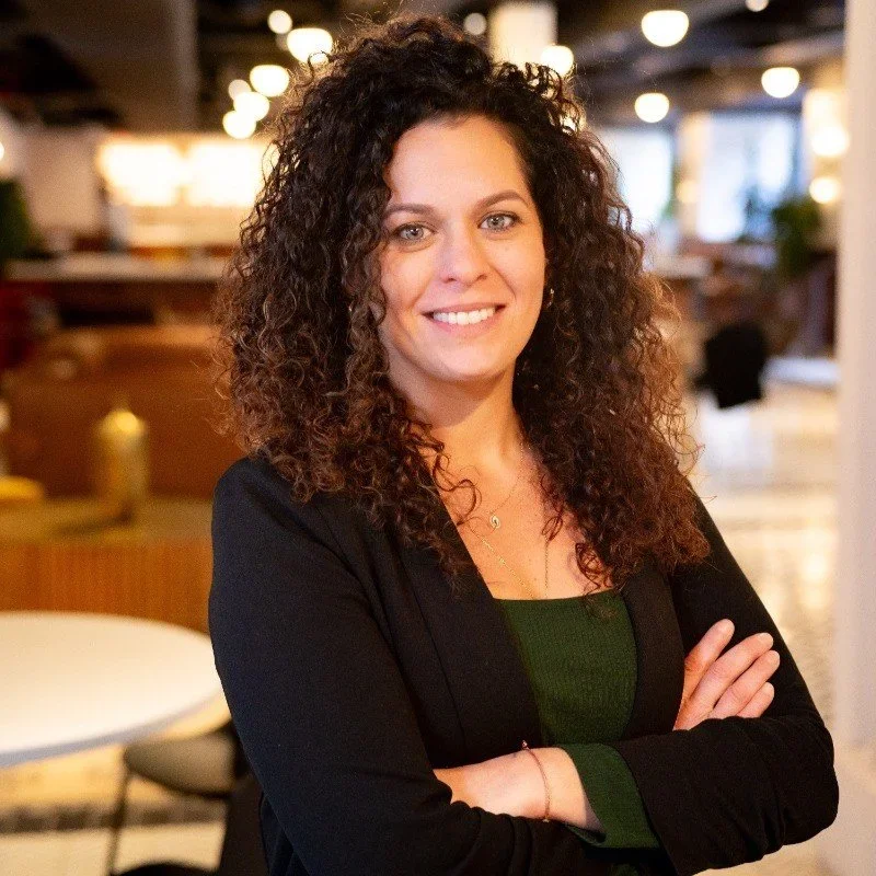 A woman with curly brown hair smiling and crossing her arms, standing in a modern, well-lit indoor space.
