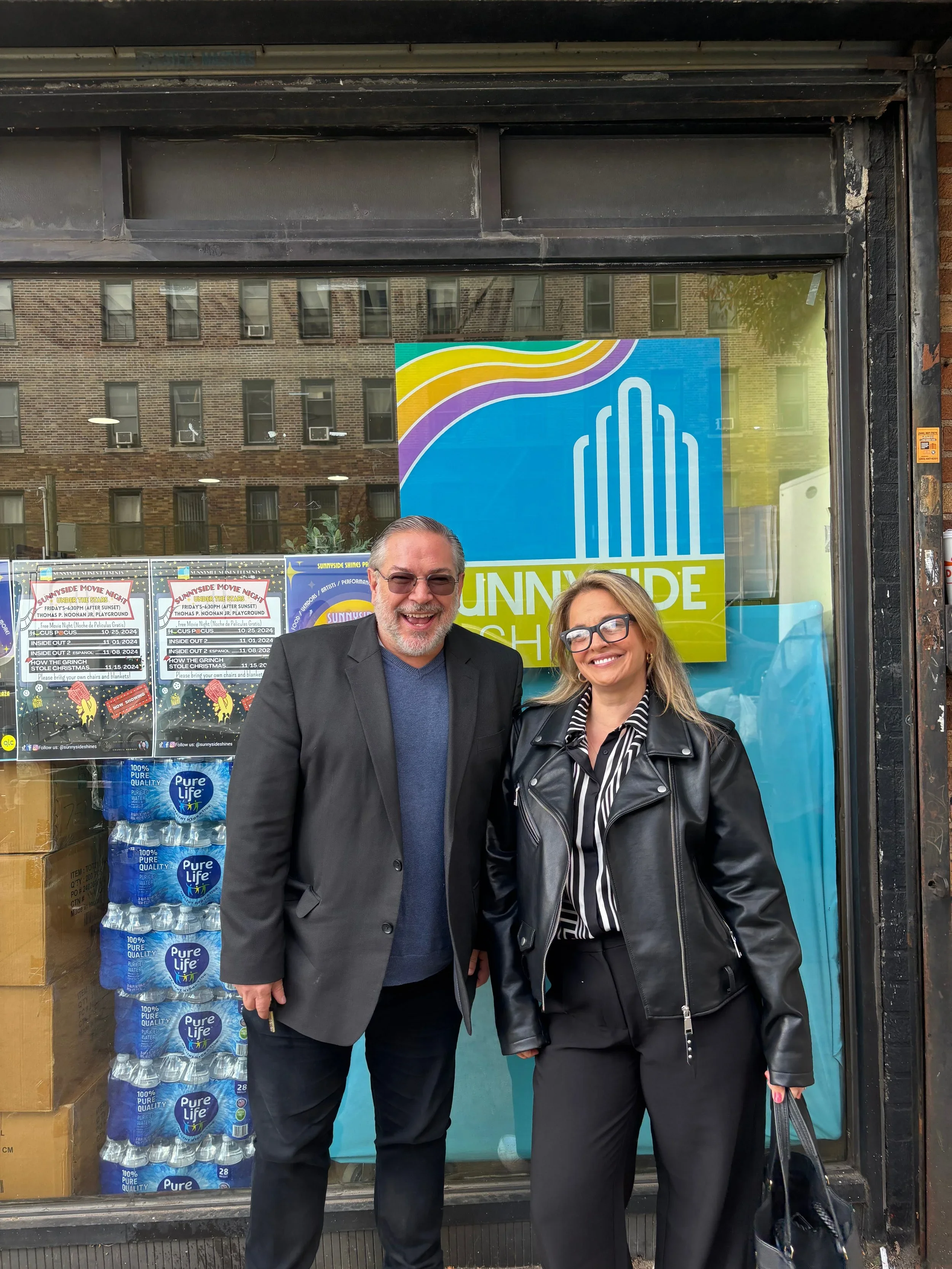 A man and a woman standing outside a storefront, smiling at the camera. The man is wearing glasses and a dark suit jacket, the woman is wearing glasses and a black leather jacket. There are water bottles and posters behind them in the store window.