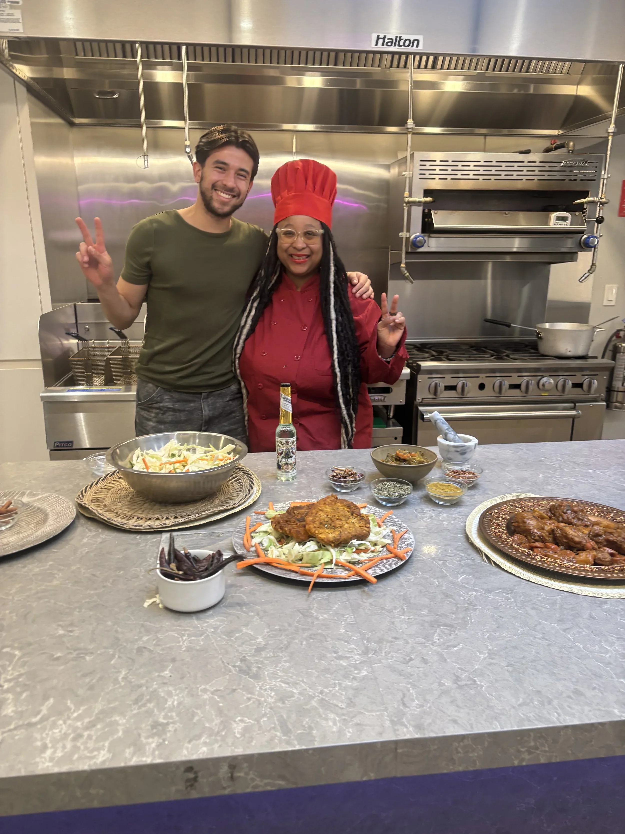Two people are standing in a commercial kitchen, smiling and making peace signs. The person on the right is wearing a red chef's hat and red outfit. On the kitchen counter in front of them, there are various dishes including fried chicken on a bed of vegetables, a bowl of soup, and other small bowls of spices or seasonings.