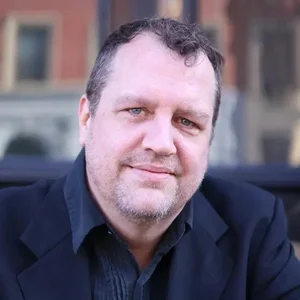 Portrait of a middle-aged man with short dark hair and a beard, wearing a dark blazer and shirt, smiling outdoors with a blurred city background.