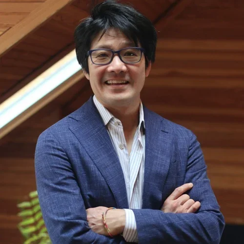 A smiling man with black hair and glasses wearing a blue suit jacket and white shirt, standing indoors near a wooden wall.