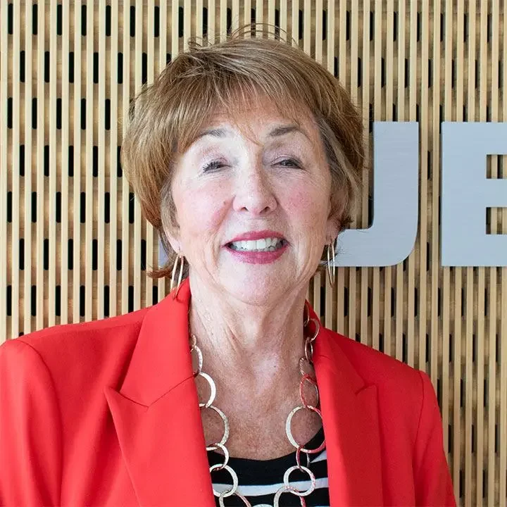 An older woman with short light brown hair, wearing a bright red blazer, a black and white striped shirt, and a silver circular necklace, standing in front of a wooden slat wall with large white letters partially visible.