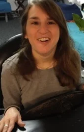 Image Description: A young white woman with long reddish-brown hair smiles at the camera, her hand extended on the surface in front of her.