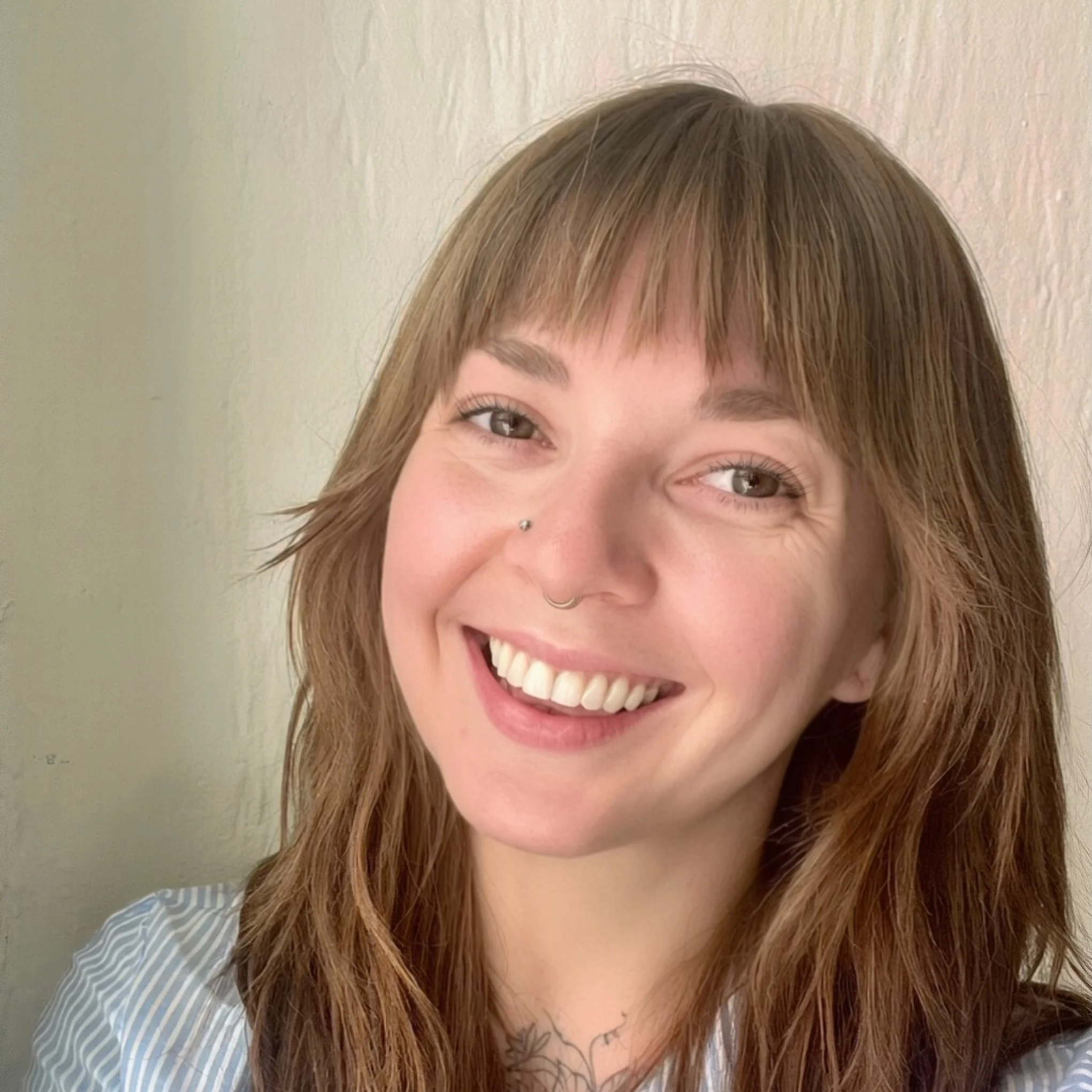 A woman with long hair and visible piercings and tattoos smiles warmly at the camera
