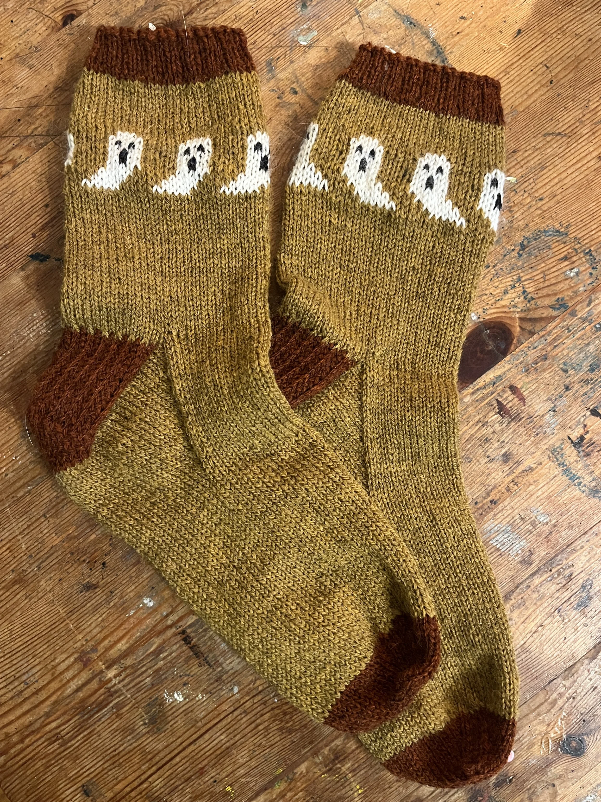 Hand-knit mustard yellow socks with little white ghosts flying around the cuffs and dark red accents.