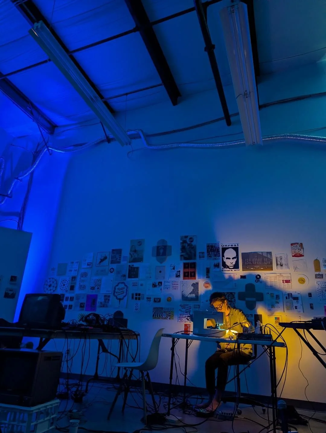 I am operating a sewing machine with a mixer and bits of paper around, with a lightbulb projecting my shadow onto the wall behind. TI am a 30-something white person with very short hair, wearing plain performance clothes and focused on my instrument.