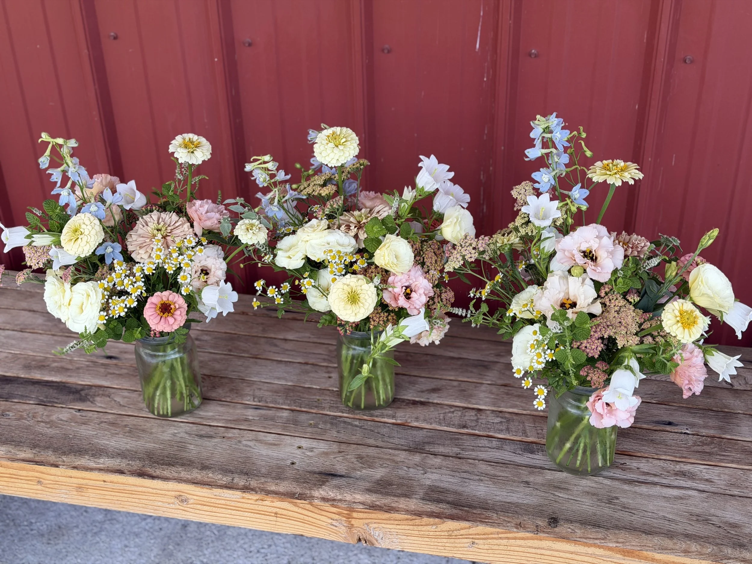 A glass jar filled with a colorful bouquet of pink, white, and orange flowers and green leaves, resting on a white wooden bench outdoors.