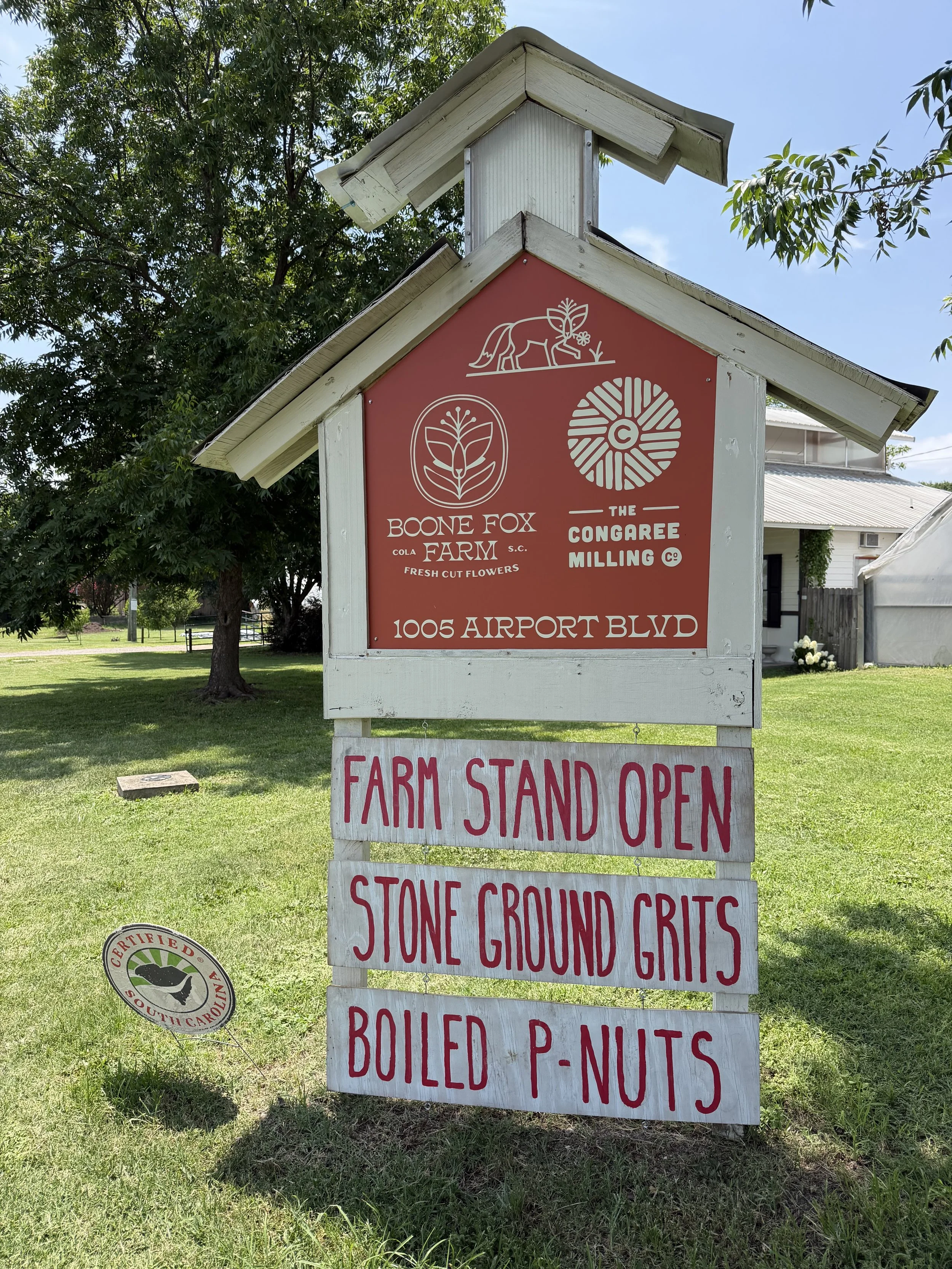 A man with a beard and mustache wearing a black t-shirt and gray overalls standing on green grass next to a signboard at a farm. The signboard reads 'City Roots Organic Farm' with an address below, and a smaller white sign with red letters says 'Farm Stand Open' followed by 'Stone Ground Grits' and 'Boiled P-Nuts.' There are trees and a white greenhouse in the background under a partly cloudy sky.