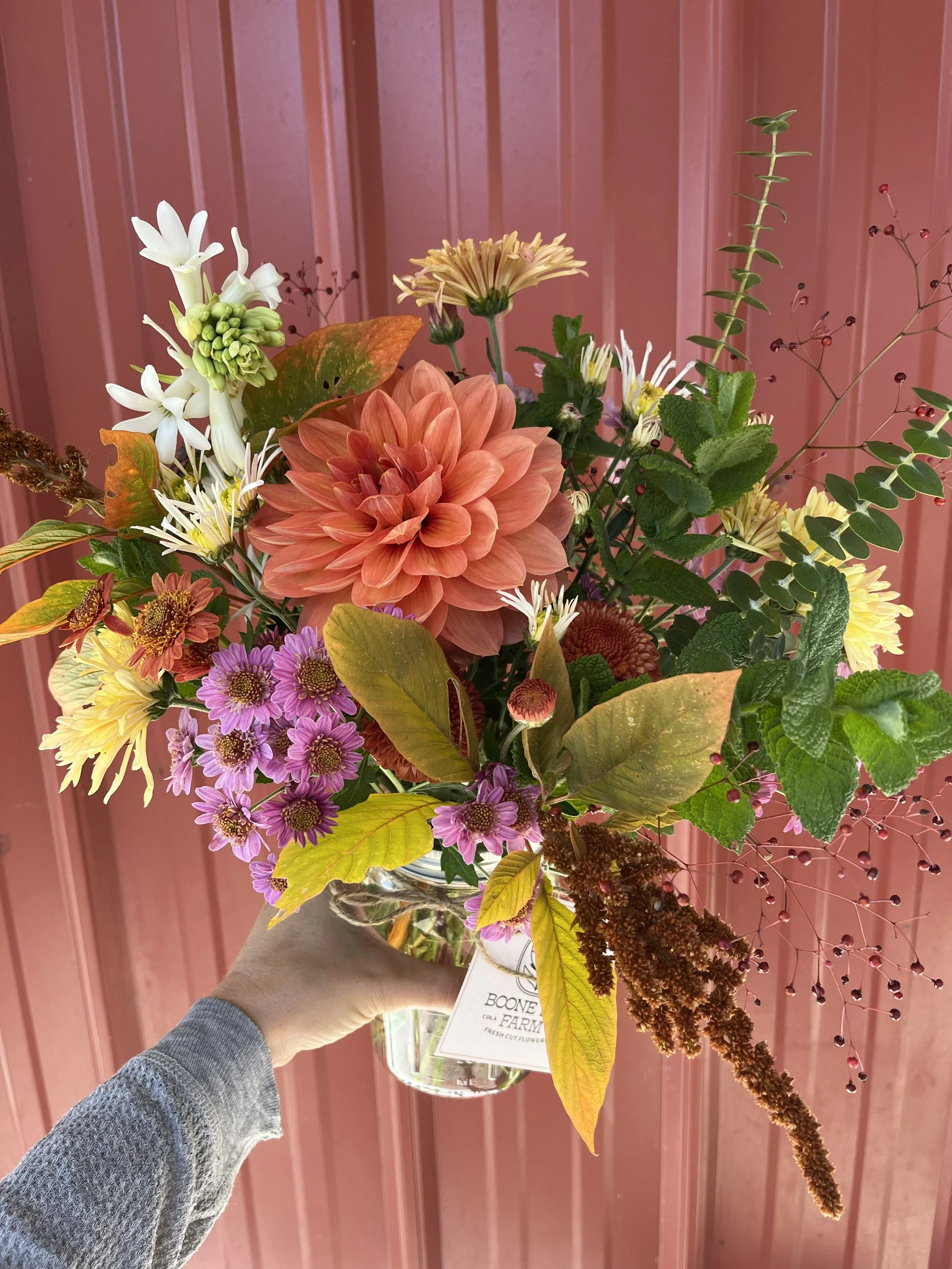 Hand holding a clear glass vase with locally grown flowers bouquet against a pink wooden background.