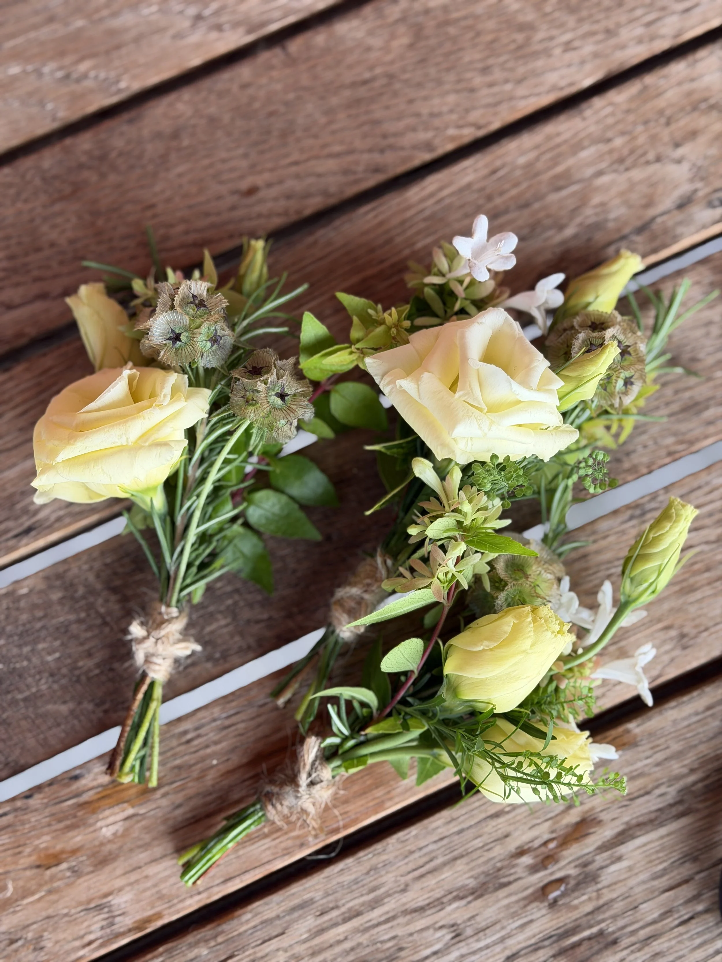 A white tray holding several boutonnieres from Boone Fox Farm made of white and purple flowers with green leaves, each tied with brown twine. Some paper notes inside the tray indicate they are for groomsmen and an extra boutonniere.