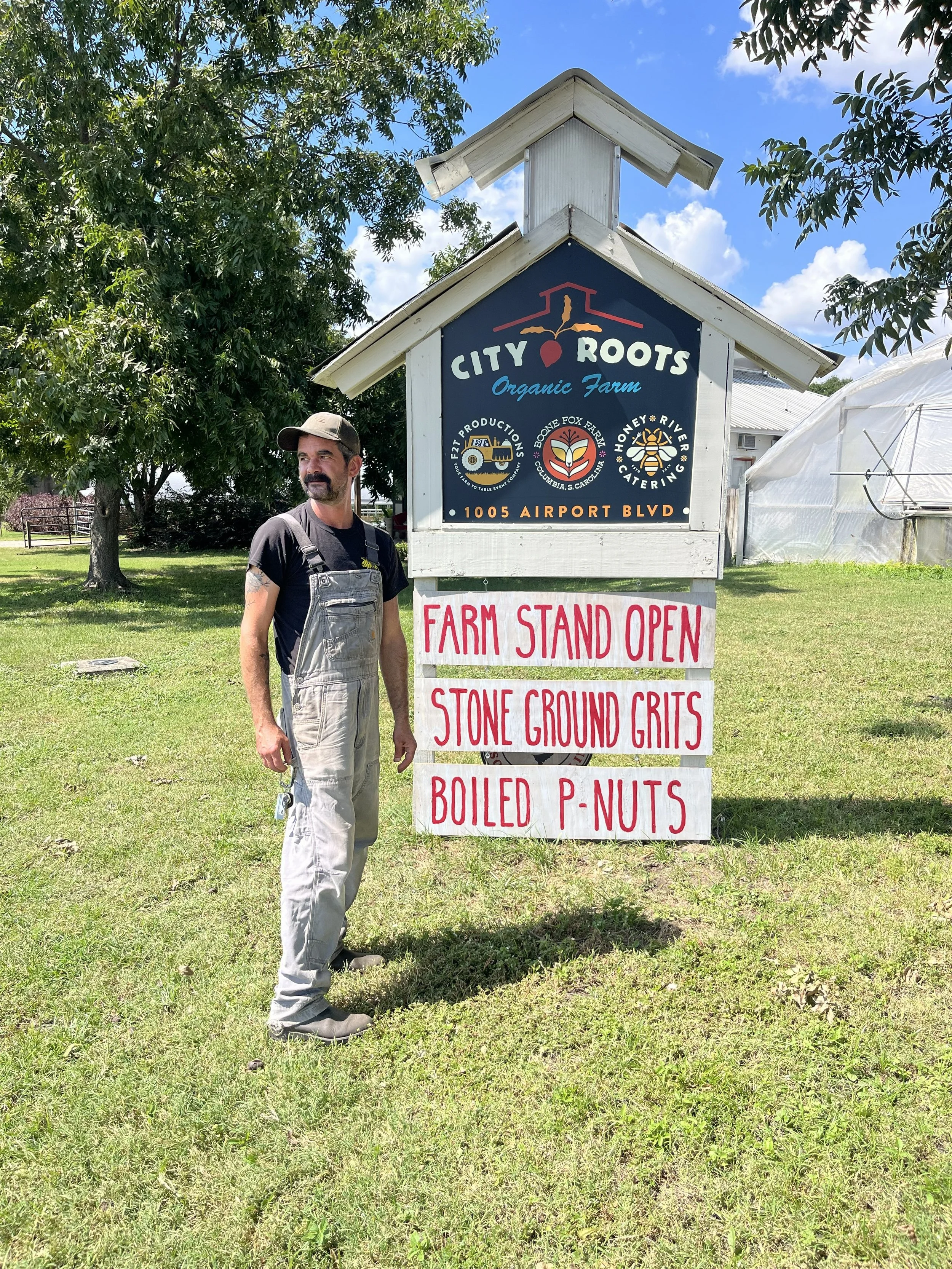A man with a beard and mustache wearing a black t-shirt and gray overalls standing on green grass next to a signboard at a farm. The signboard reads 'City Roots Organic Farm' with an address below, and a smaller white sign with red letters says 'Farm Stand Open' followed by 'Stone Ground Grits' and 'Boiled P-Nuts.' There are trees and a white greenhouse in the background under a partly cloudy sky.