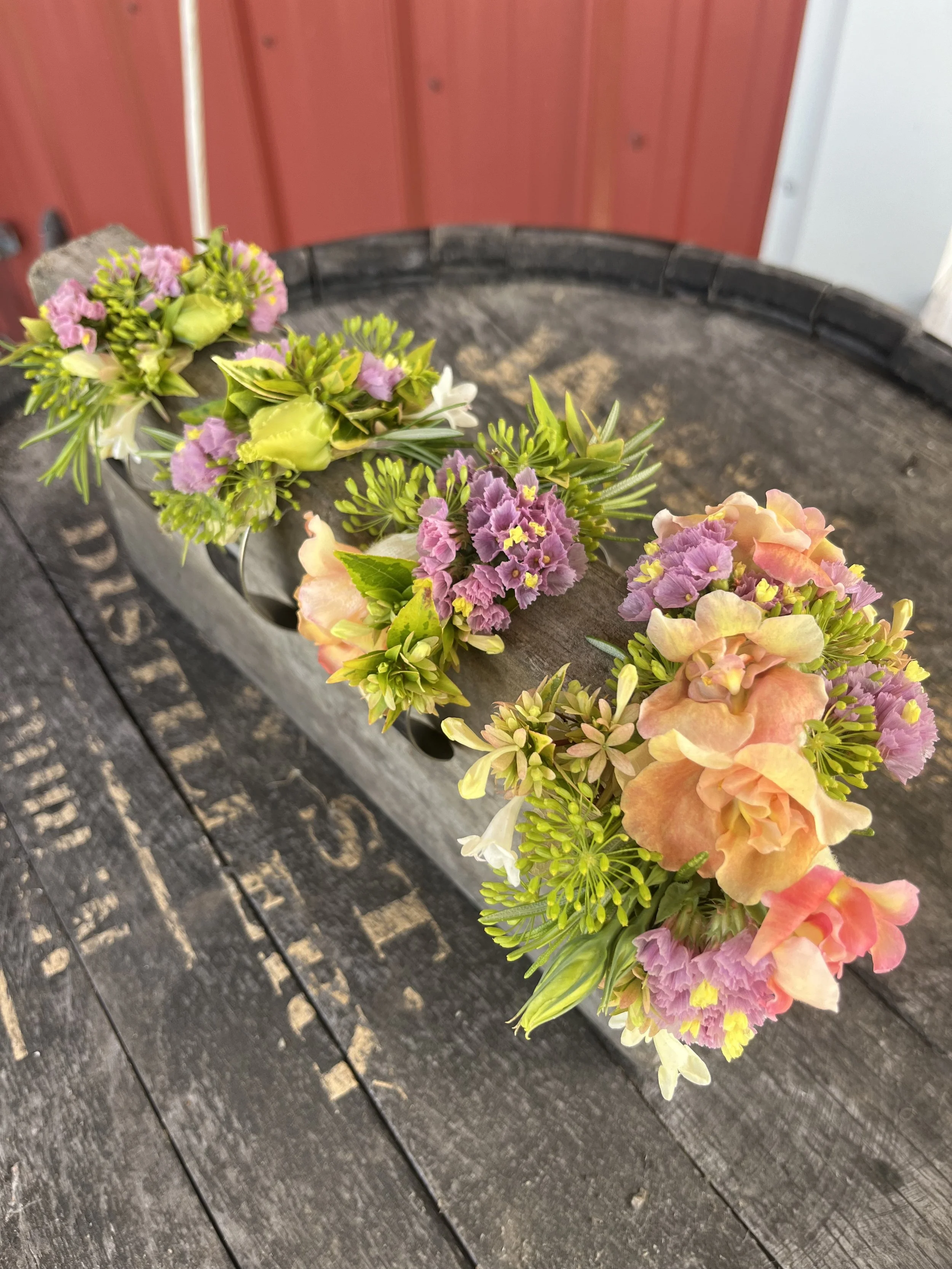 Person holding a small bouquet of white, yellow, and purple flowers with green leaves and rosemary sprigs against a light-colored textured wall.