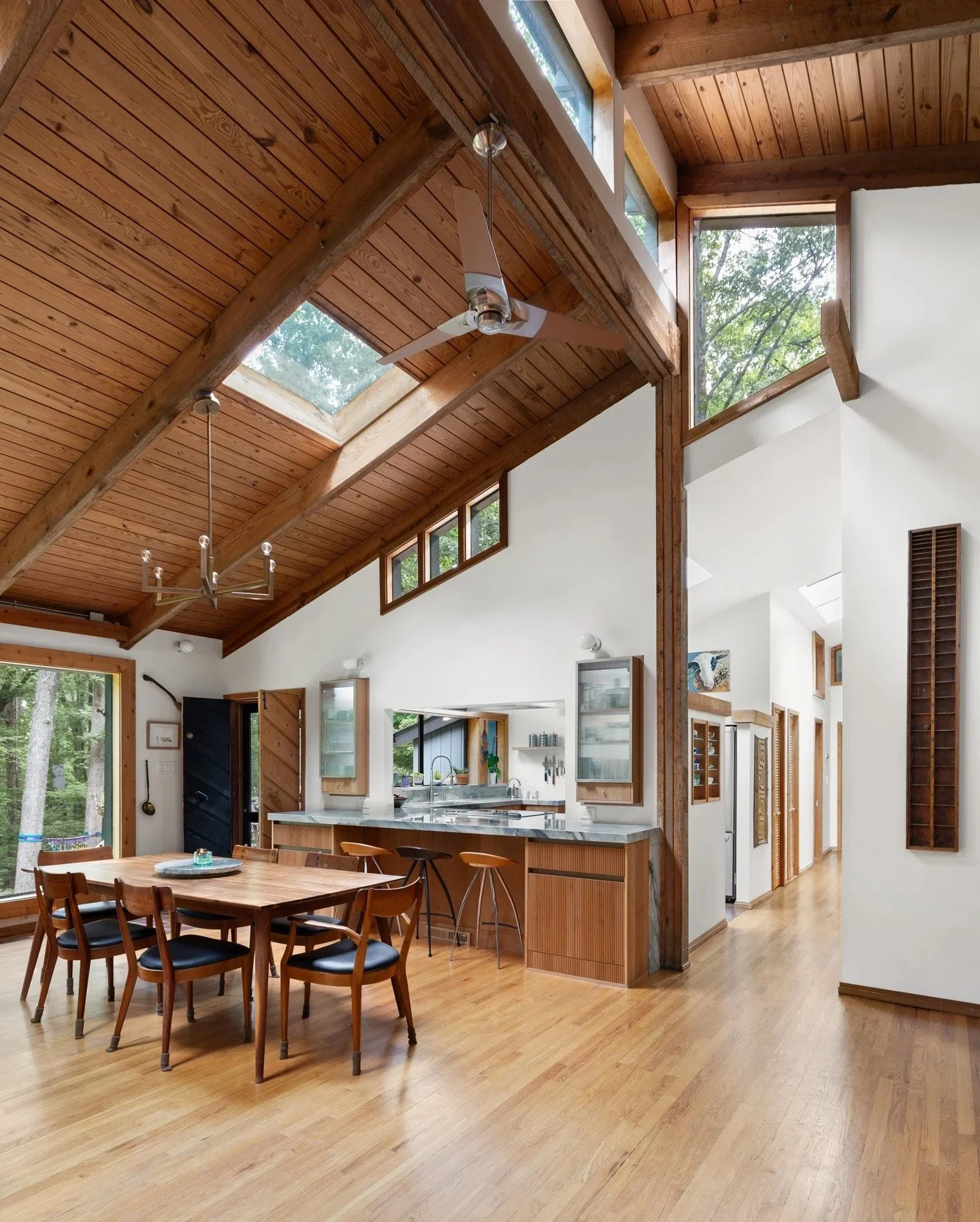 A mid-century great room with vaulted wood ceilings and wood floors. Large windows and skylights let natural light in. A modern kitchen with wood cabinets and custom built ins updates is in the background.