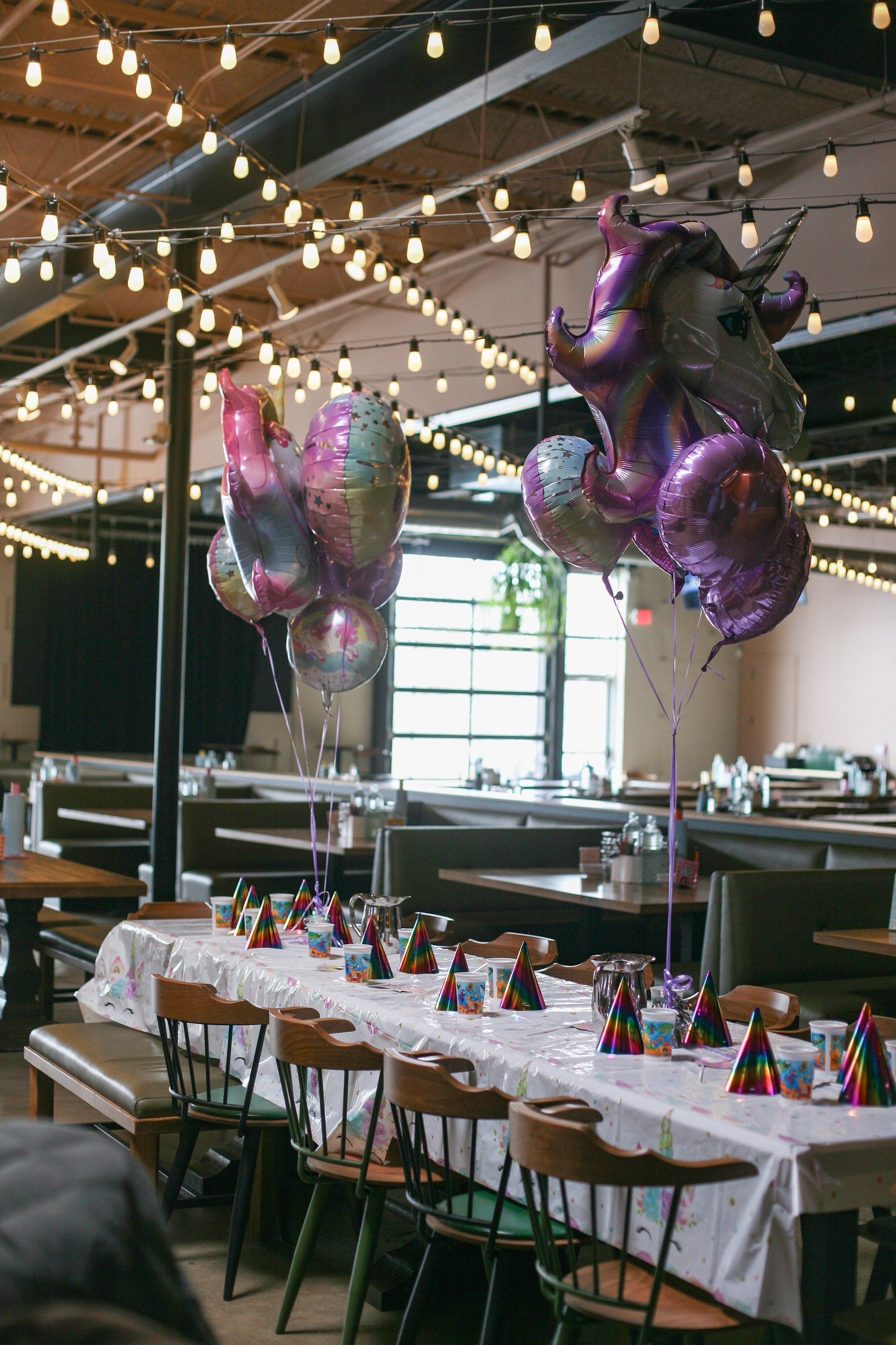 Indoor party setup with a long table covered in a white tablecloth with colorful party hats, cups, and plates. Balloons and string lights are hanging from the ceiling, creating a festive atmosphere.
