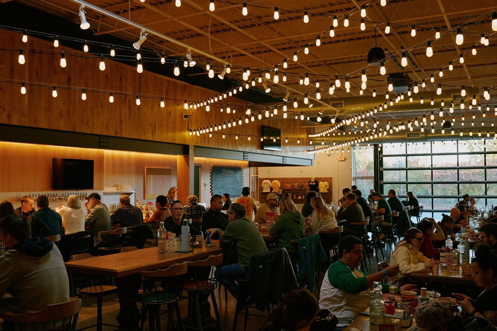 A busy indoor restaurant filled with people dining. The ceiling is decorated with numerous hanging string lights. There are large windows in the background letting in natural light.