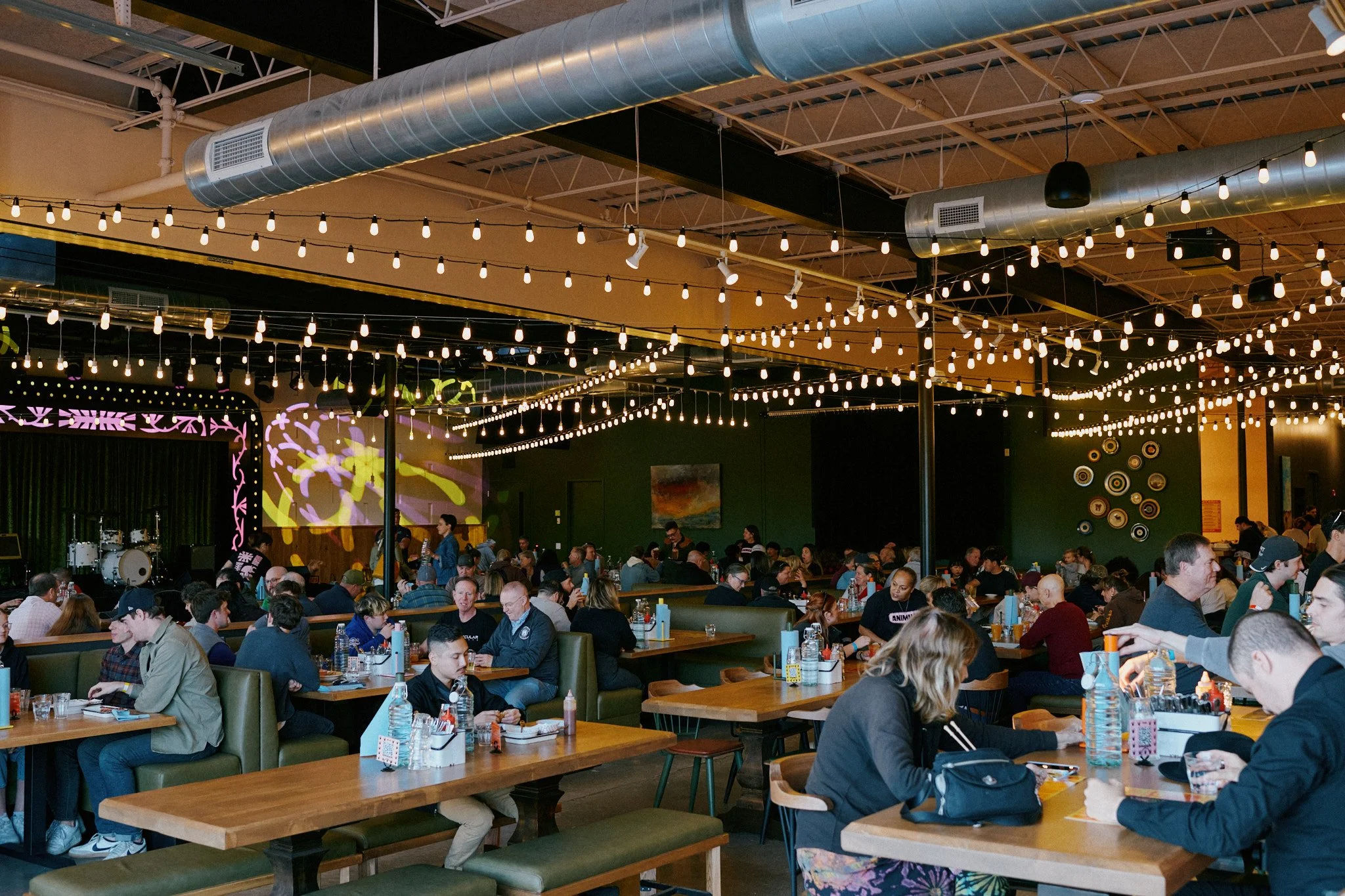 Indoor restaurant filled with people sitting at tables, eating and talking. Decor includes string lights hanging from the ceiling and wall art.