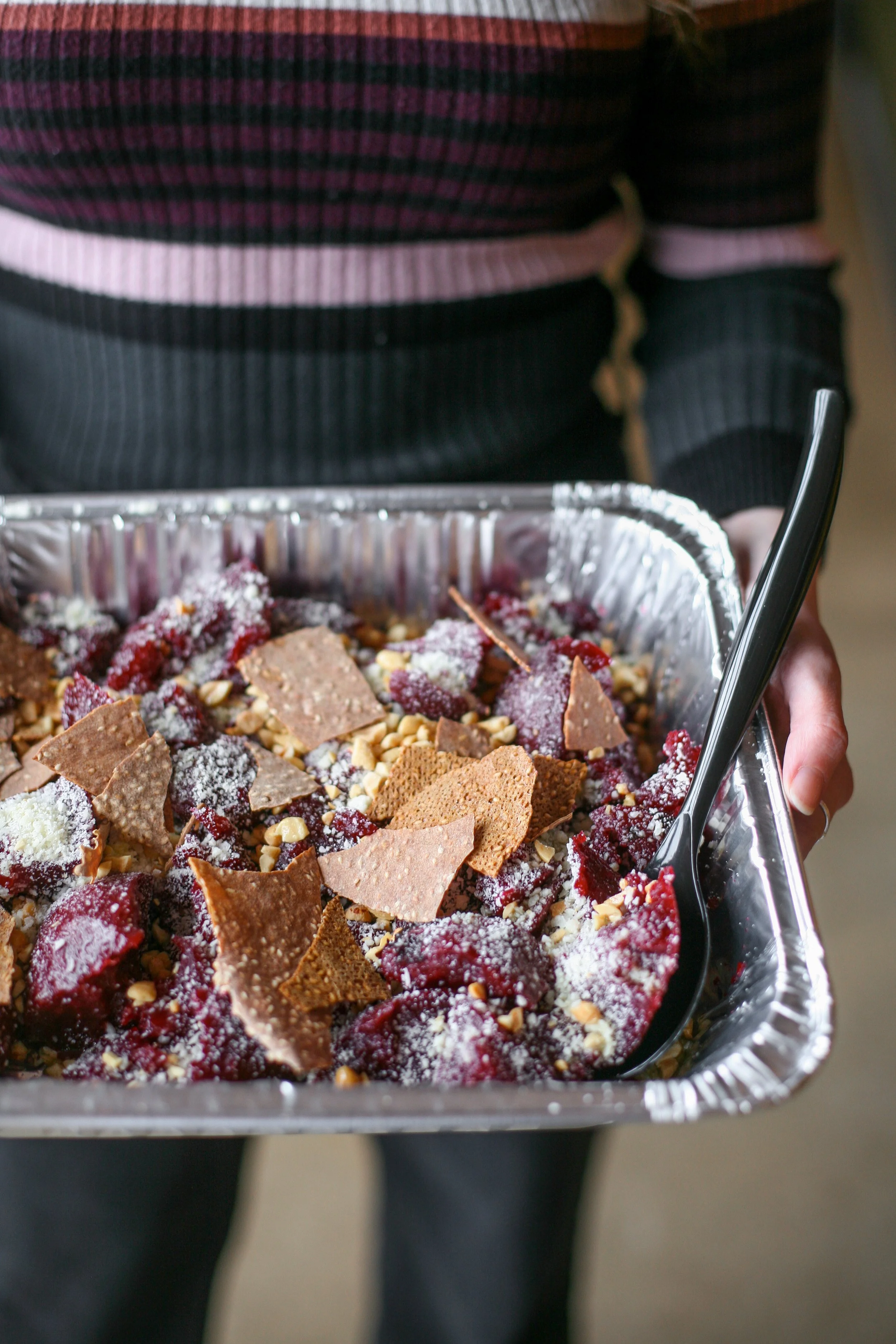 Person holding a foil tray filled with a pickled beet salad topped with benne seed crackers, cotija, and olive oil. This dish is available for catering.