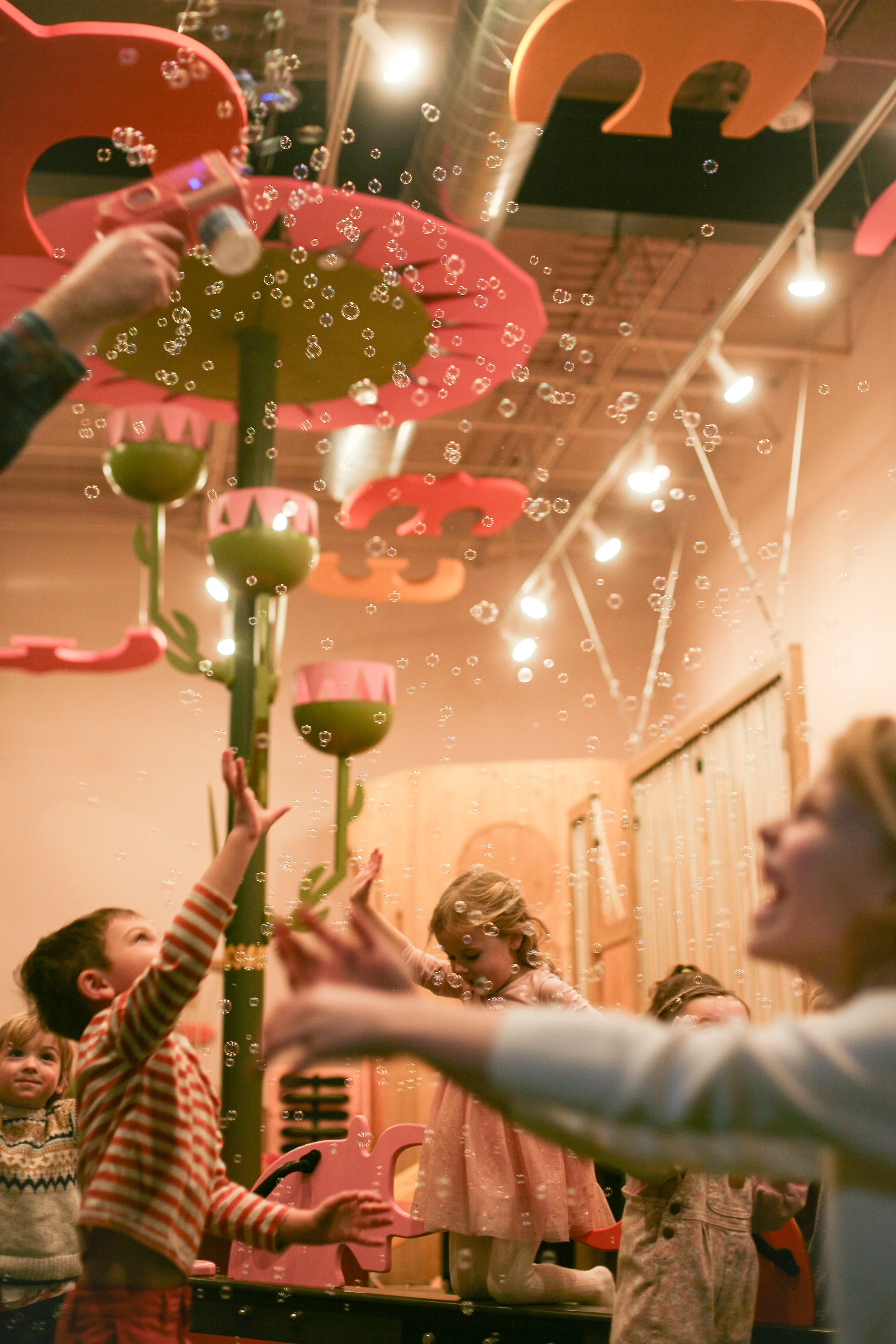 Children celebrating a birthday party, blowing bubbles, and playing with a pink and green pinwheel on a table, with pink and orange balloons hanging from the ceiling.