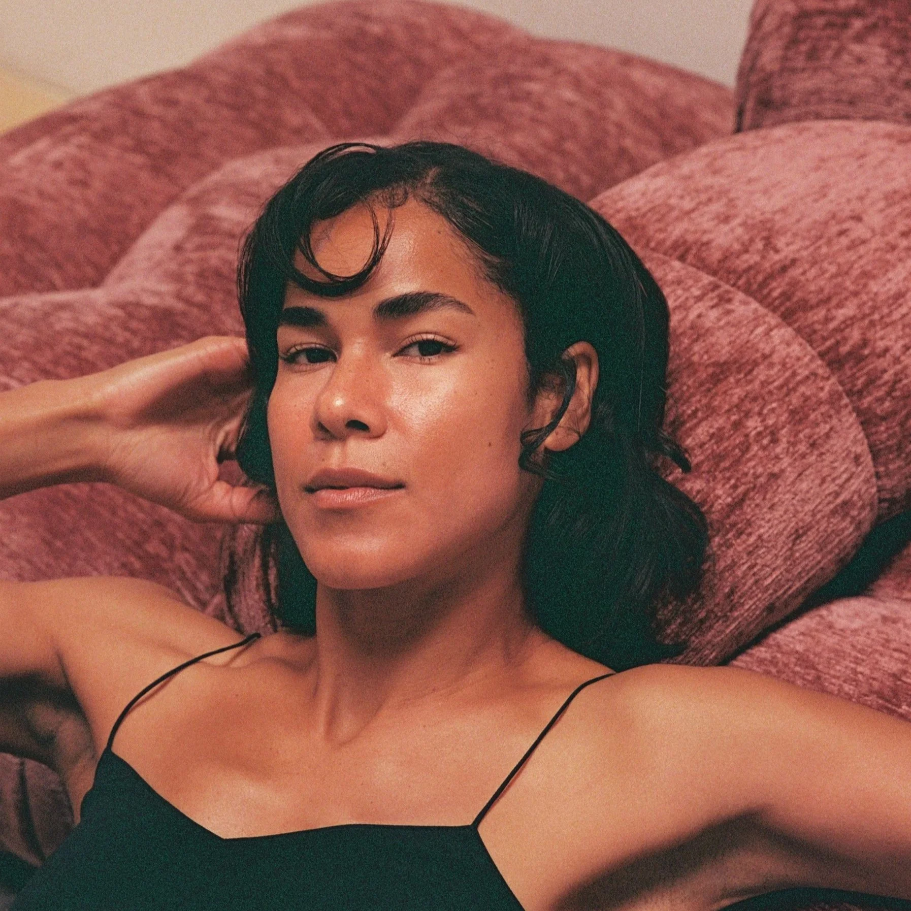 A woman with dark hair lying on a pink, textured, fluffy blanket, wearing a black spaghetti strap top, with a relaxed and confident expression.