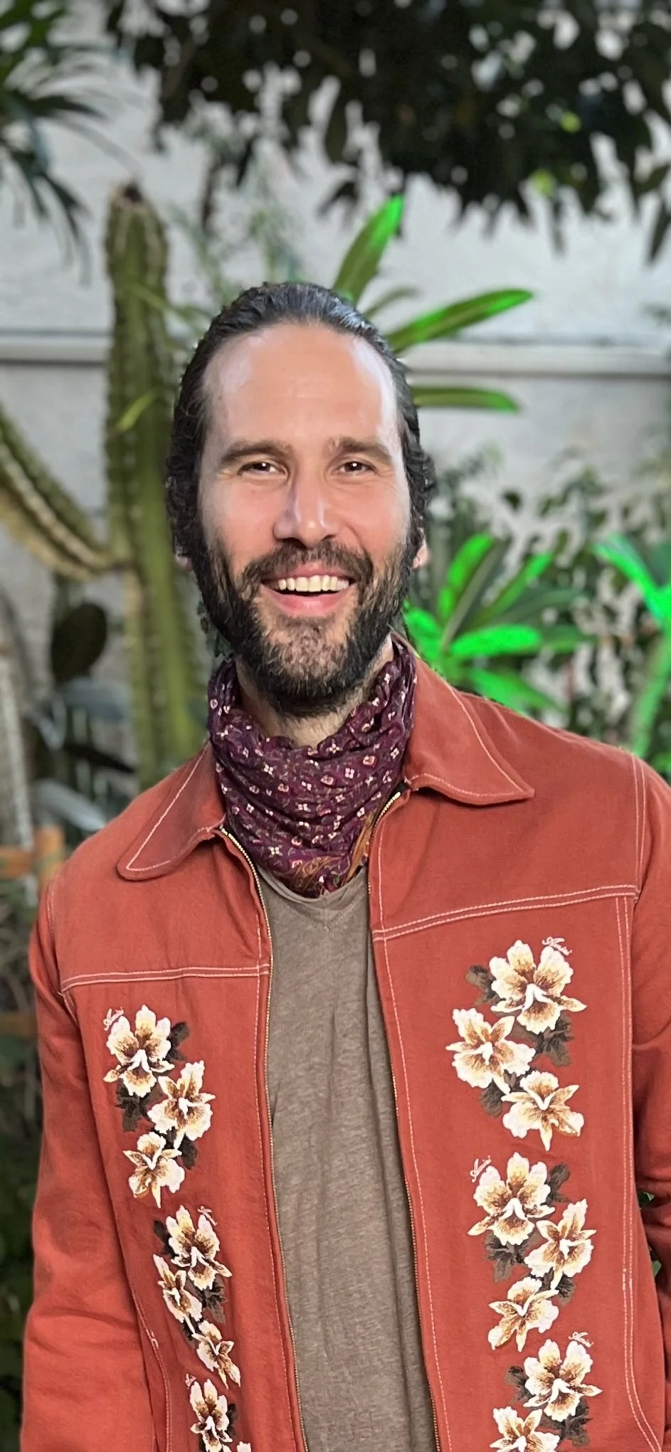 Owner of Furaha Artists and Furaha Sound, Jan Christiansen, with dark hair and a beard, wearing an orange jacket with floral embroidery, a gray shirt, and a purple bandana, standing in front of green plants.
