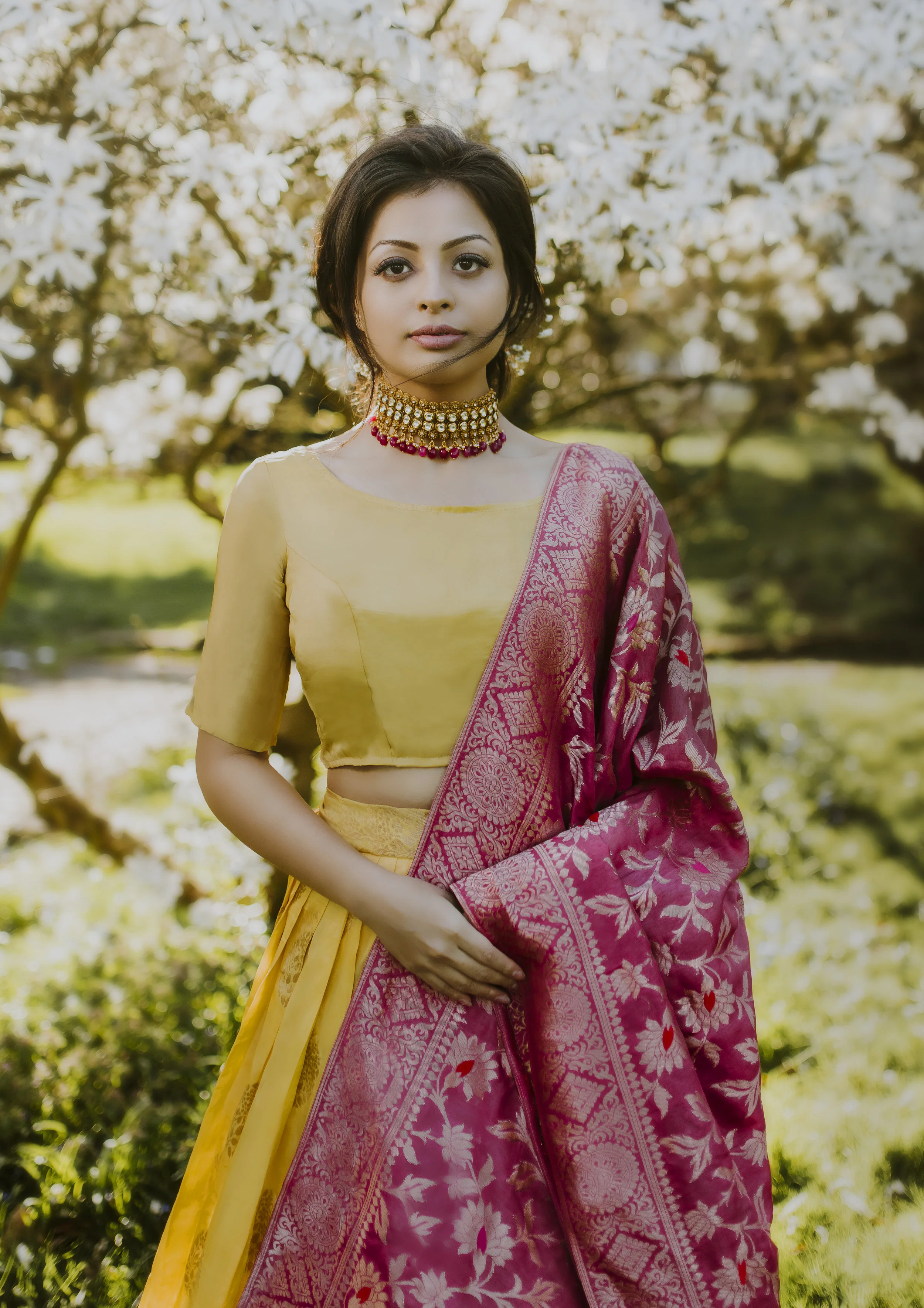 A woman standing outdoors in front of flowering trees, wearing a yellow and pink traditional Indian outfit with a choker necklace.