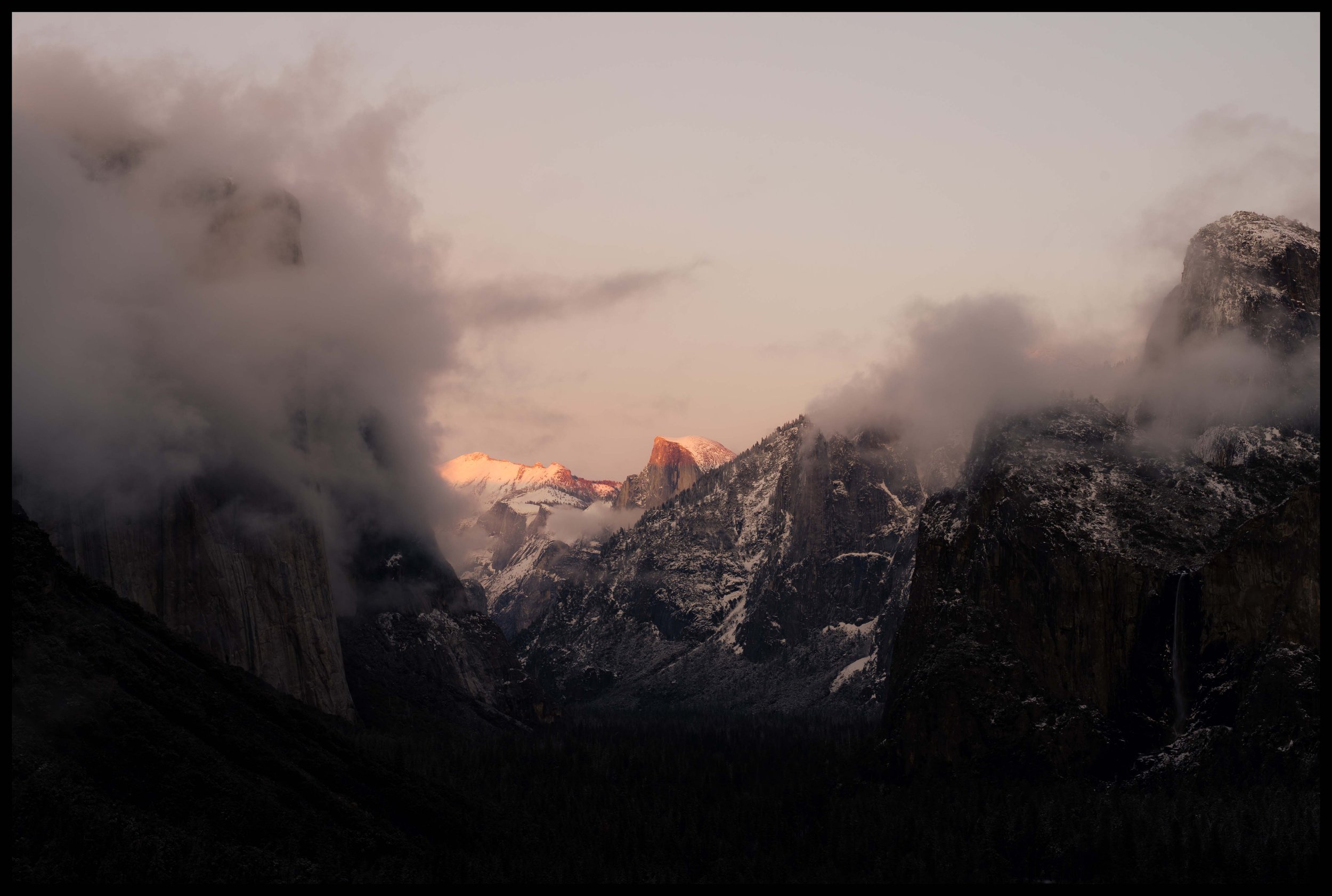 The last light of day illuminates a peak in Yosemite while other cliffs are obscured by low-hanging clouds.