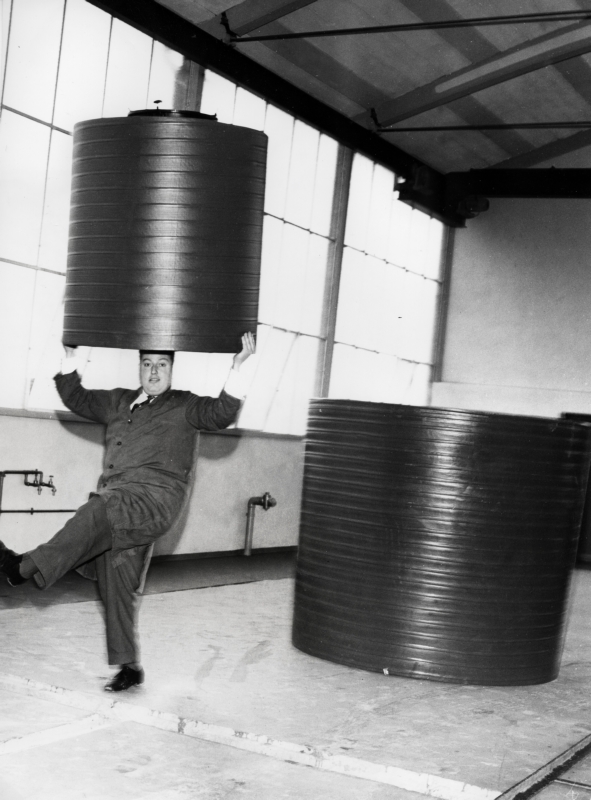 Een werknemer - gekleed in een stofjas - balanceert een kunststof vat (polyetheen) op zijn hoofd. A man balancing a plastic (polyethene) container.
