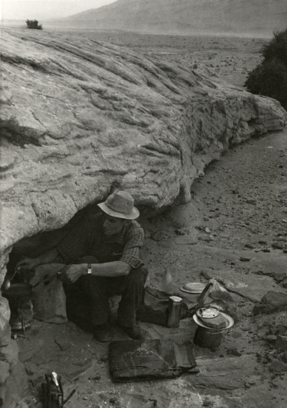 Om zich te beschermen tegen de straffe Sahara wind kookt Jan Glissenaar op zijn primus in een rotsspelonk. Algerije, 1953. To protect himself against the strong Sahara wind Jan Glissenaar is cooking a meal on a primus in a cavern. Algeria, 1953.