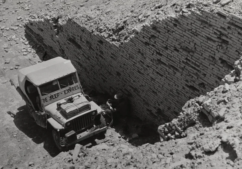 In de stad van aartsvader Abraham rust de Jeep-Expres uit in de schaduw van de torentempel van de Ziggoerat. Jan Glissenaar leunt tegen de tempelmuur. Irak, Ur der Chaldeeen, 1954. In the city of patriarch Abraham the Jeep Expres is taking a rest at the feet of the Great Ziggurat of Ur of the Chaldees. Jan Glissenaar is leaning against the wall of the temple. Iraq, 1954.