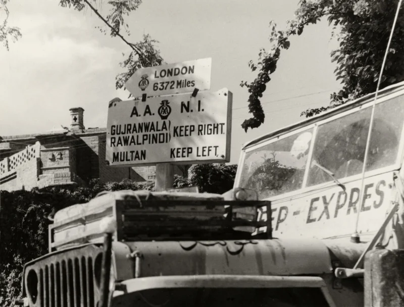 De jeep passeert een verkeersbord in Lahore met de afstand in mijlen naar Londen. Het tweetal is op de terugweg. Pakistan, Lahore, 1955. The jeep passing a traffic sign in Lahore with the distance in miles to London. The two travellers are on their way back. Pakistan, Lahore, 1955.
