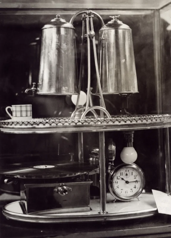 Een tableau om te gebruiken bij het ontwaken. Bovenop bevinden zich twee ketels met een kraantje, waaruit de koffie getapt kan worden. Onderop een grammofoon en een wekker. Parijs, ca. 1932.