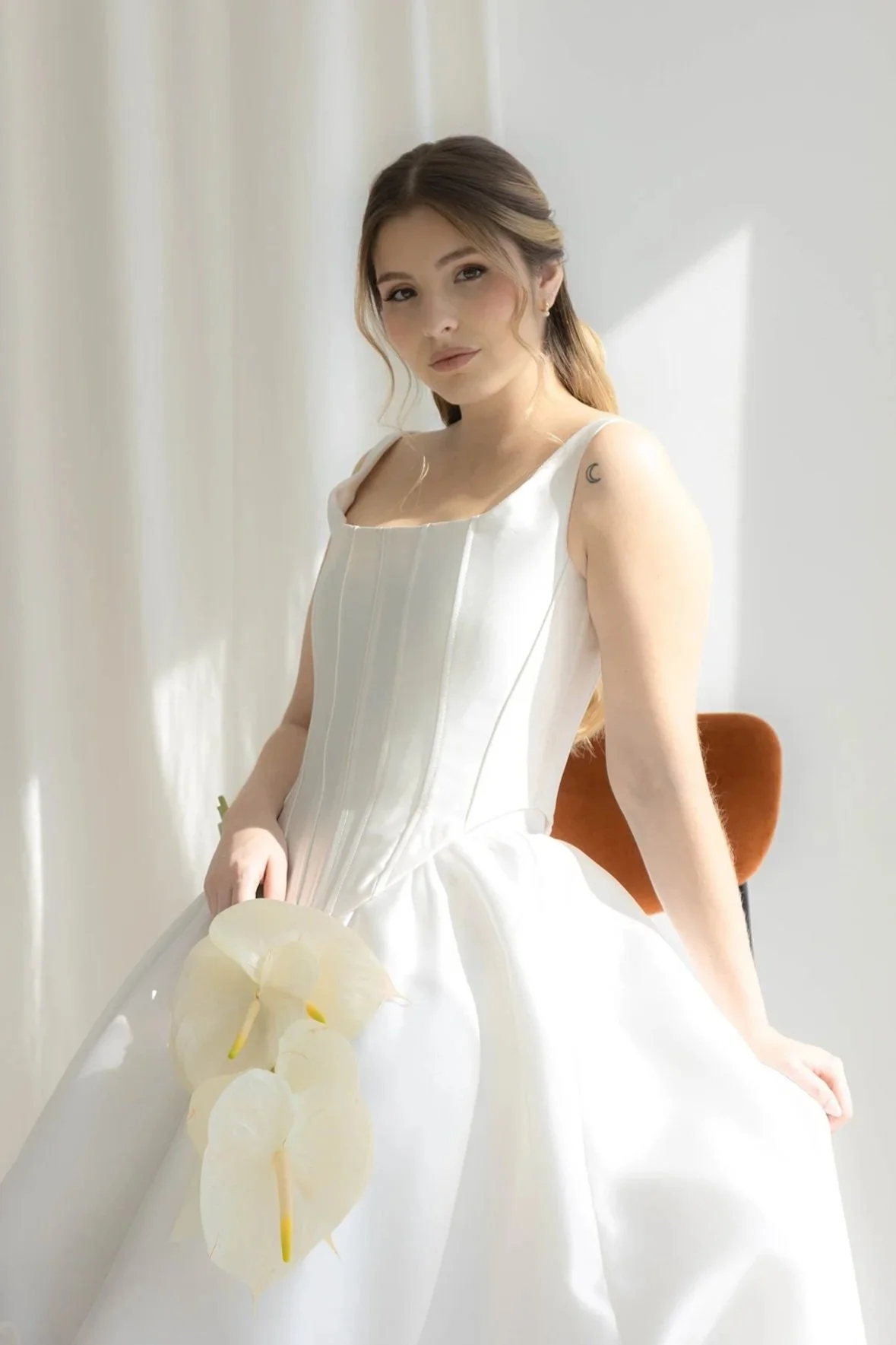 Mujer joven con vestido blanco sentada en una silla, sosteniendo un ramo de flores blancas, en un entorno con luz natural.