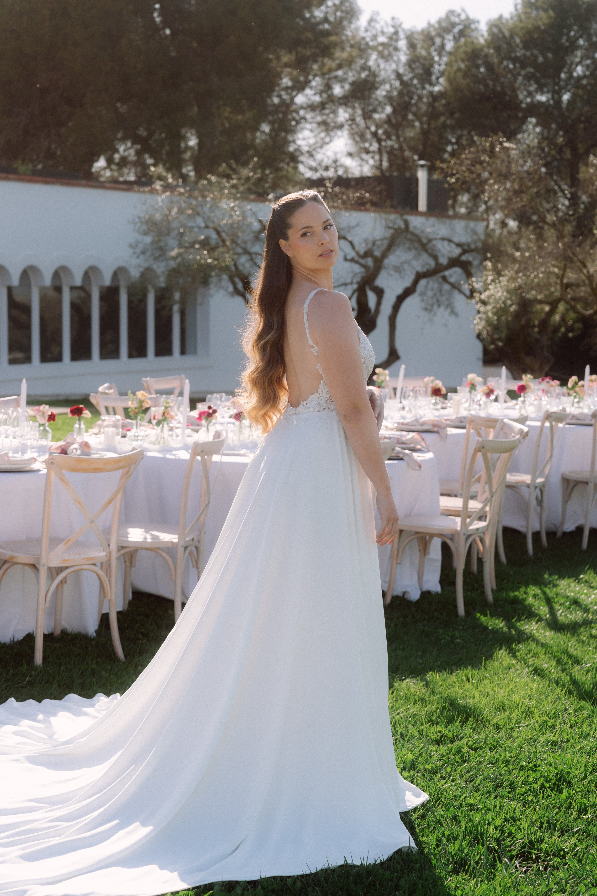 Mujer con vestido de novia en exterior, con mesa de banquete decorada con flores y velas al fondo, durante el día.
