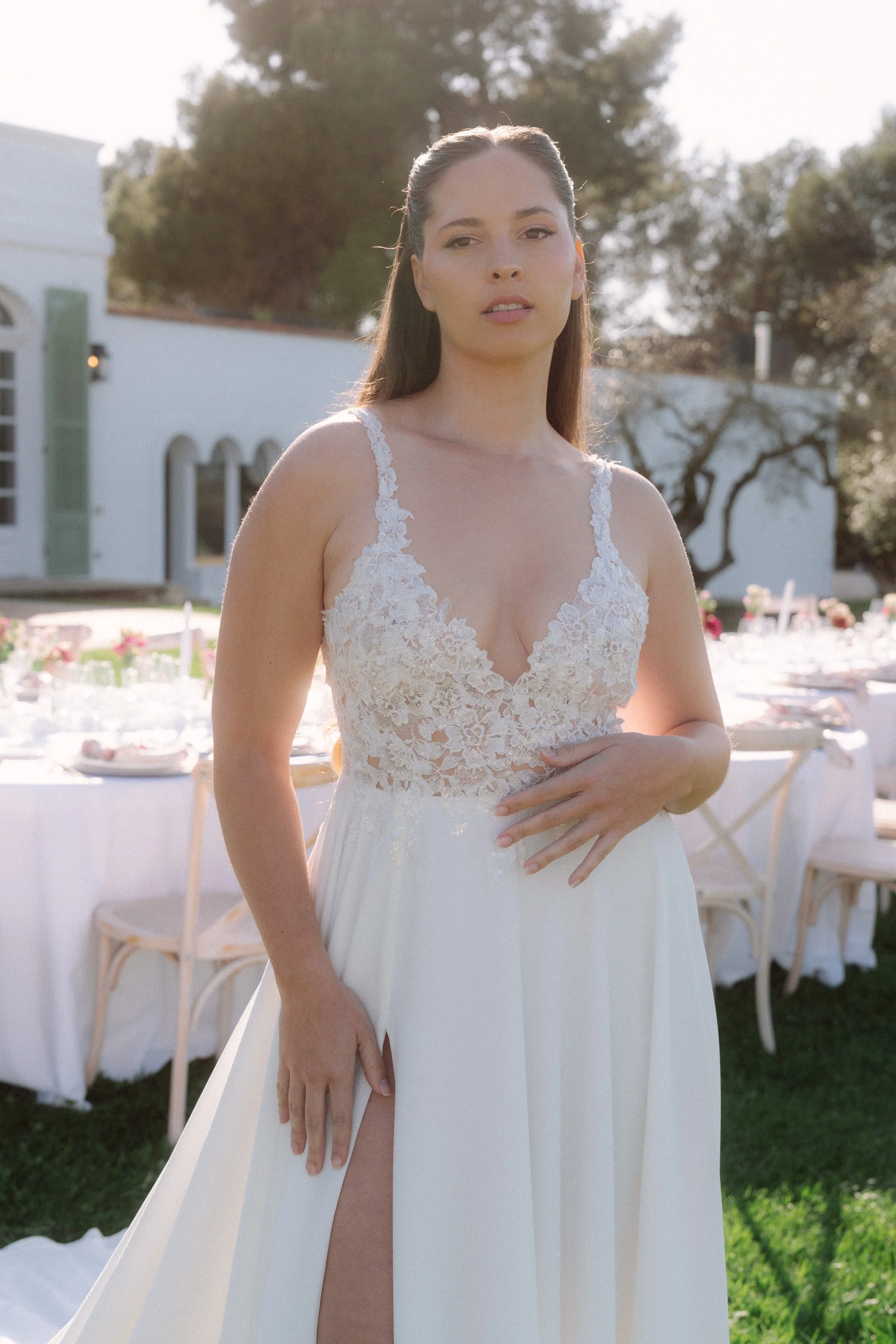Mujer con vestido de novia en un evento al aire libre, en un lugar con árboles y mesas decoradas para una celebración.