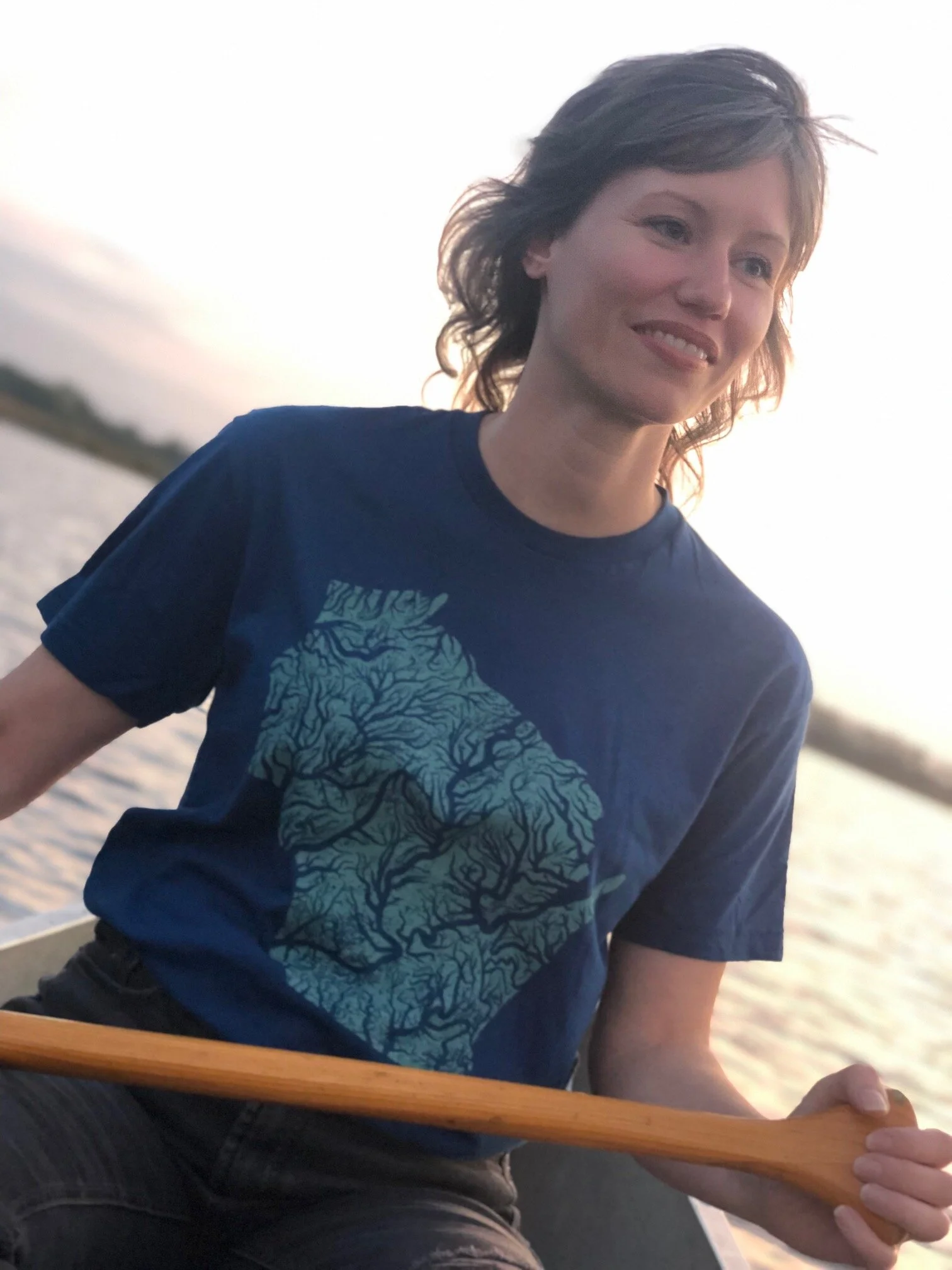 A woman wearing a deep blue graphic tshirt with a stylized representation of the Wisconsin river system screen printed in sea green on the front and the HomeLove logo on the nape.