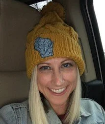 A women wearing the chunky cable knit beanie style stocking hat with pom on top.  Mustard with an embroidered Wisconsin rivers patch.