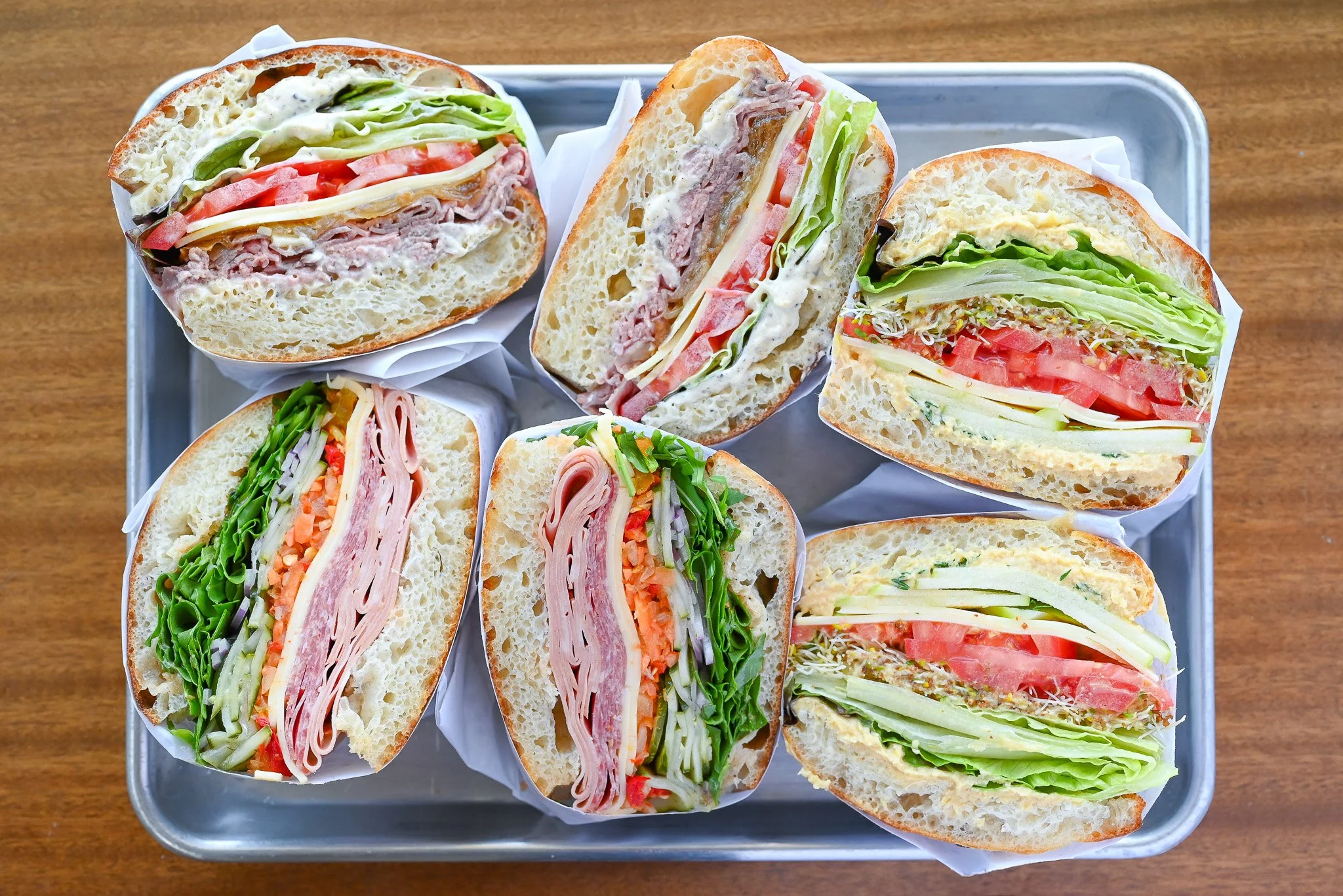 Assorted cut sandwich halves with lettuce, tomato, cheese, turkey, ham, and sprouts on a metal tray.