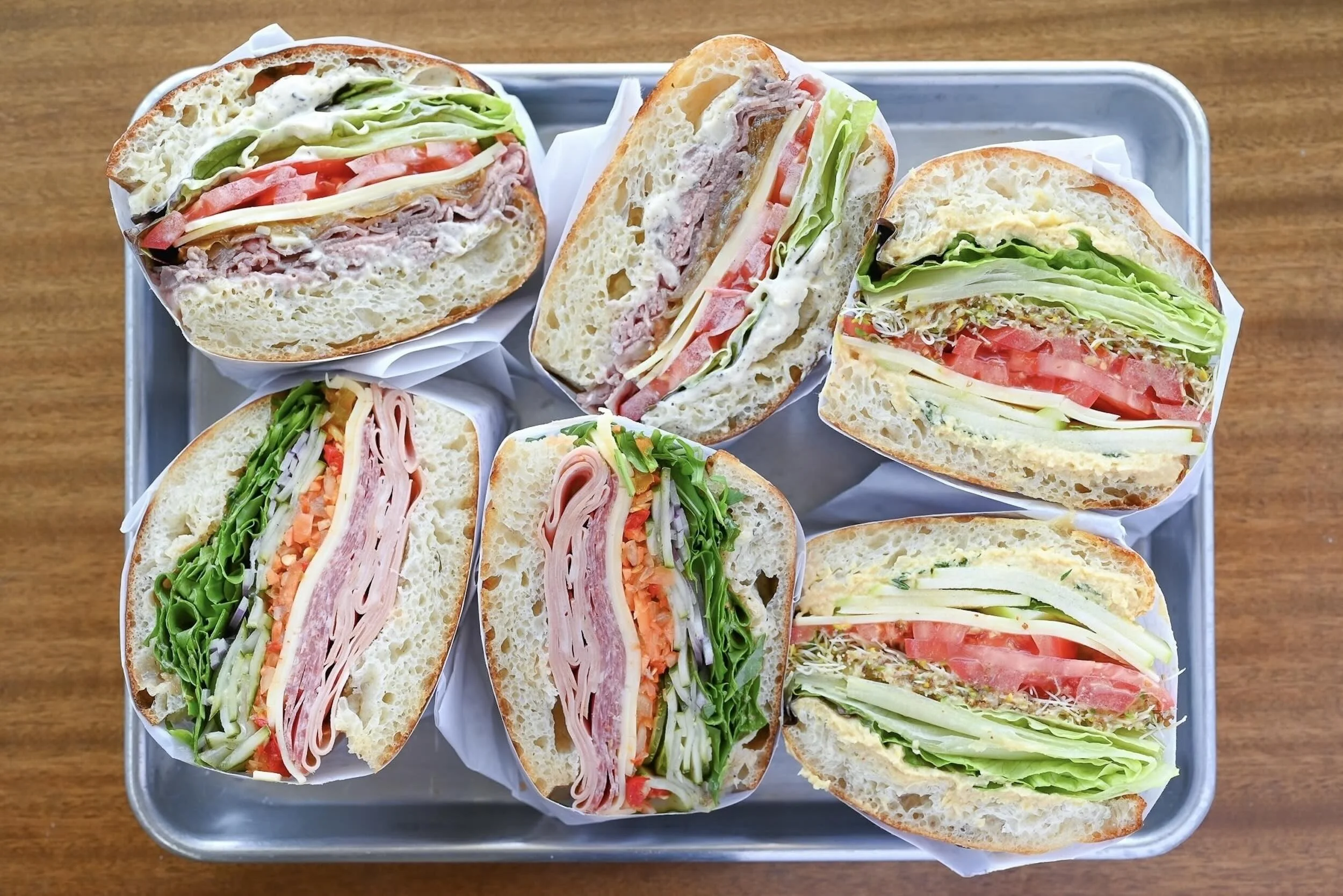 Seven assorted sandwich halves with various meats, cheeses, and vegetables on a metal tray